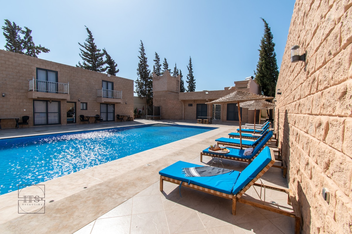 A large outdoor area features a private pool with clear blue water surrounded by sun loungers. The smooth stone patio is bordered by trees, with two bungalows providing a backdrop for a relaxing space. Shade is offered by umbrellas placed over the loungers.