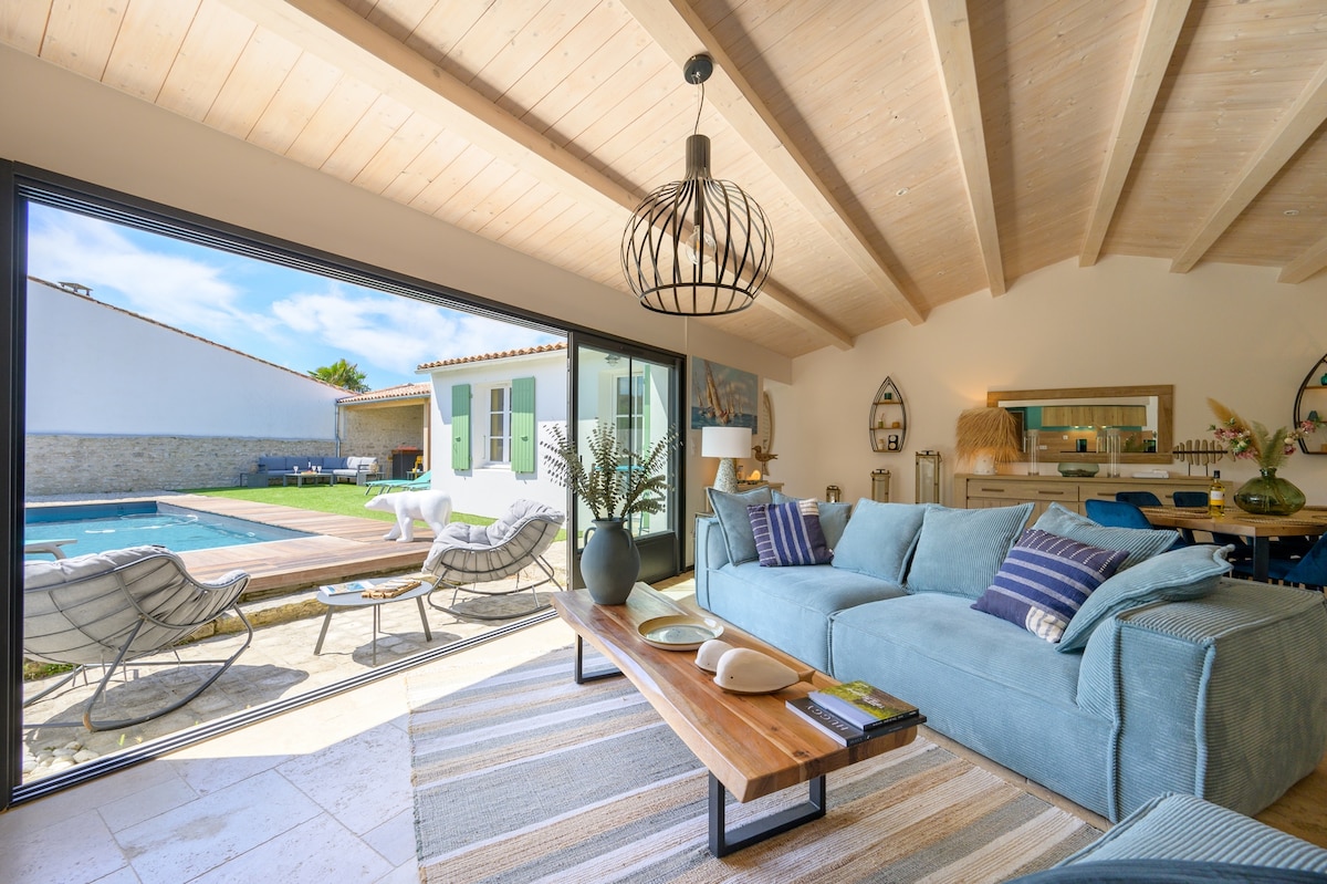 A light-filled living area features large glass doors opening to the poolside. A comfortable blue sofa and wooden coffee table create a welcoming space. A stylish light fixture hangs from the wooden ceiling, while outdoor seating is visible near the pool, enhancing the tranquil atmosphere.