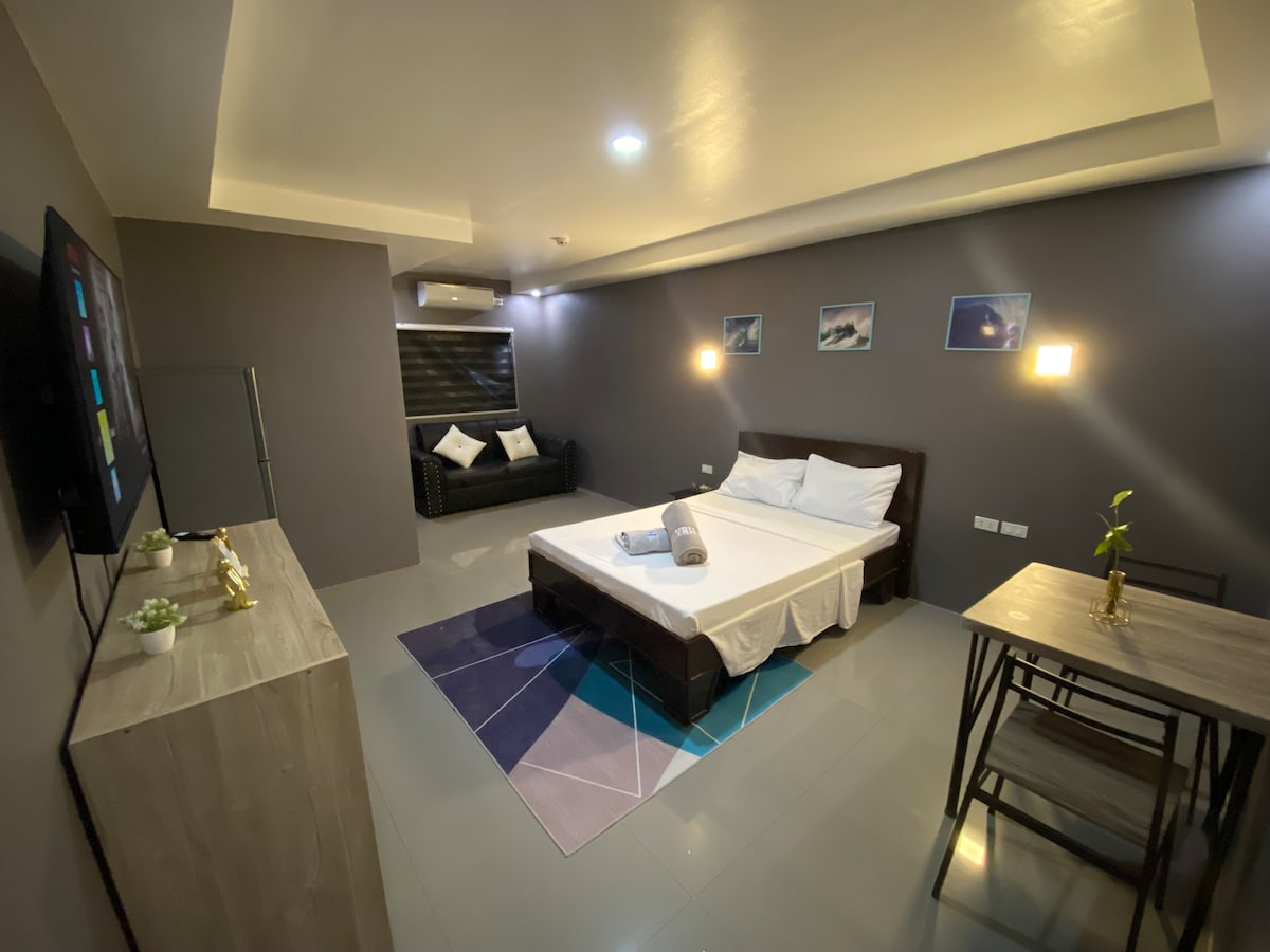 A modern room features a spacious bed with fresh linens and a rolled towel placed in the center. A relaxed seating area includes a dark couch, and a dining table with two chairs is positioned nearby. Soft lighting enhances the neutral grey walls.