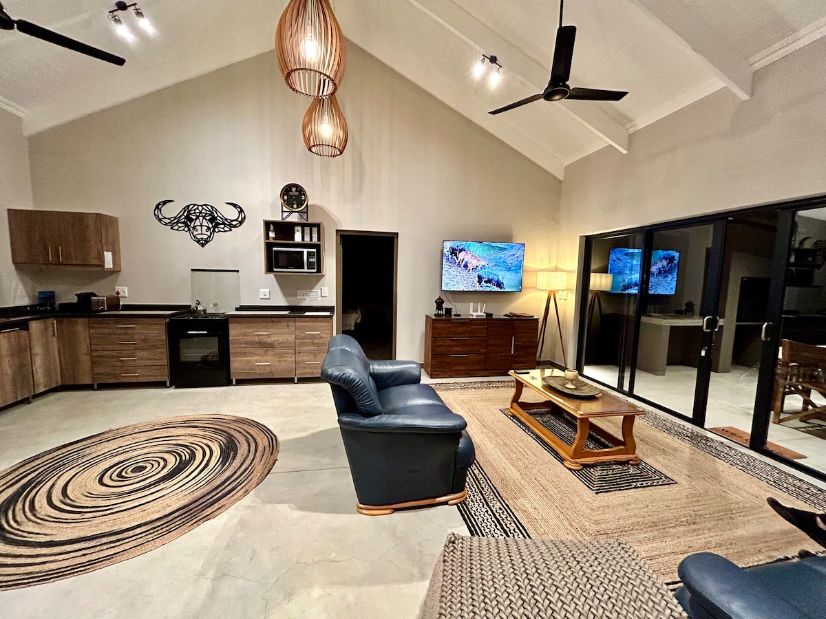 An open-plan living space features a modern kitchen with wooden cabinetry and a cozy lounge area. Two ceiling fans circulate air, and a wall-mounted flat-screen TV is positioned above a stylish wooden cabinet. Sliding glass doors allow natural light to fill the room.