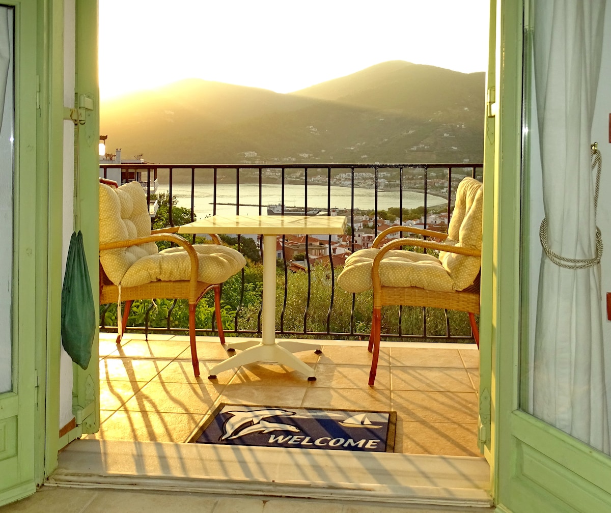 A private balcony is seen, featuring two cushioned chairs and a small table, facing the hills of Skopelos Town. A welcome mat lies at the entrance. Natural light warms the space, hinting at sunset, while lush greenery surrounds the area.
