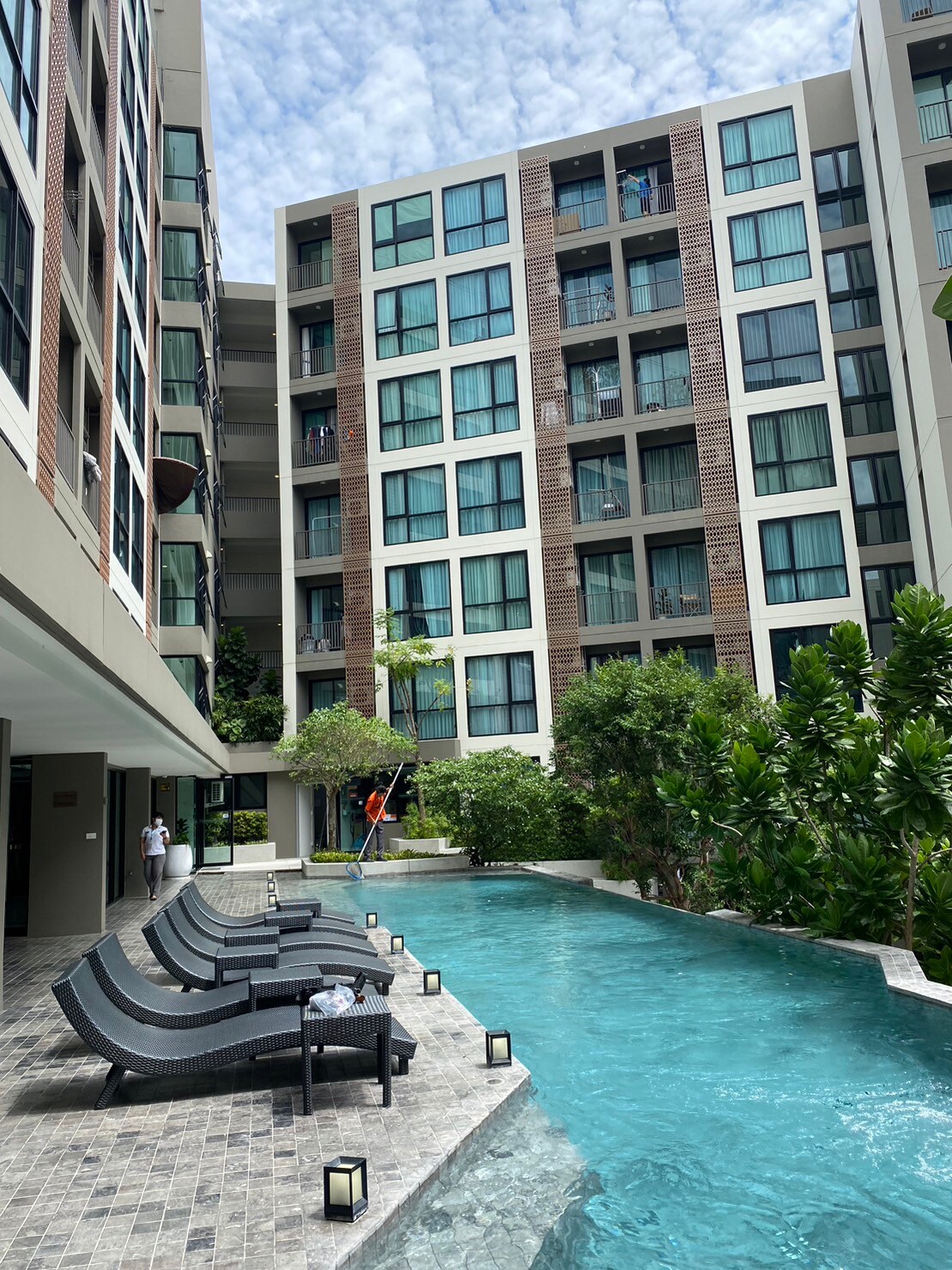 A modern apartment complex surrounds a serene swimming pool, lined with comfortable loungers. Lush greenery and flowering plants add a refreshing touch to the outdoor space, while large windows in the building reflect natural light, creating a relaxing atmosphere.