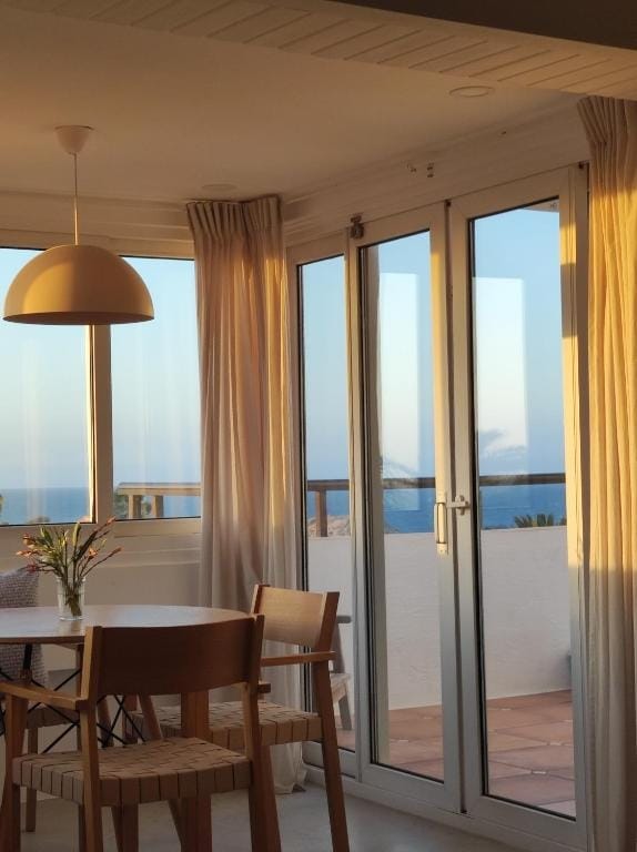 A dining area with a round table surrounded by wooden chairs is visible. Sheer curtains frame large glass doors that open to a sea view. Soft evening light filters through, creating a serene atmosphere.