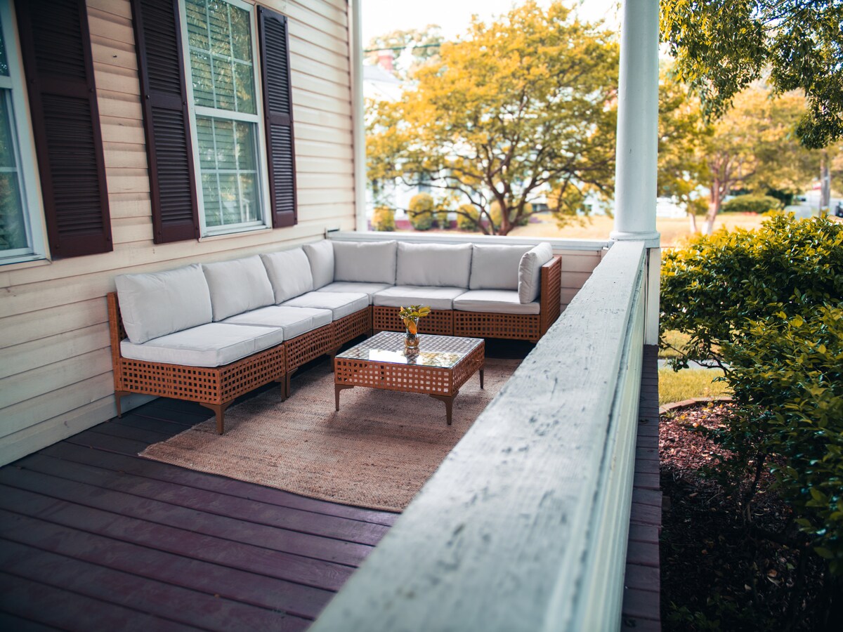 A cozy outdoor seating area is shown, featuring a sectional sofa with light-colored cushions arranged around a small coffee table. A vase with flowers serves as a centerpiece. Lush greenery and trees are visible in the background, creating a peaceful setting.