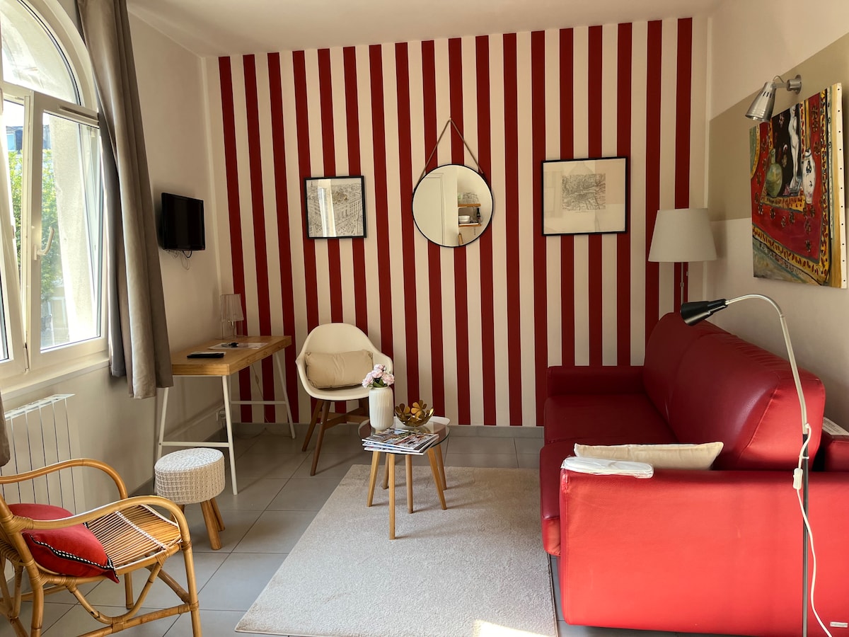The living area showcases a red striped accent wall, complemented by a red sofa and natural wooden furniture. A small round table is positioned beside the sofa, with a floor lamp offering soft lighting. A mirror and artwork adorn the walls, enhancing the cozy ambiance.