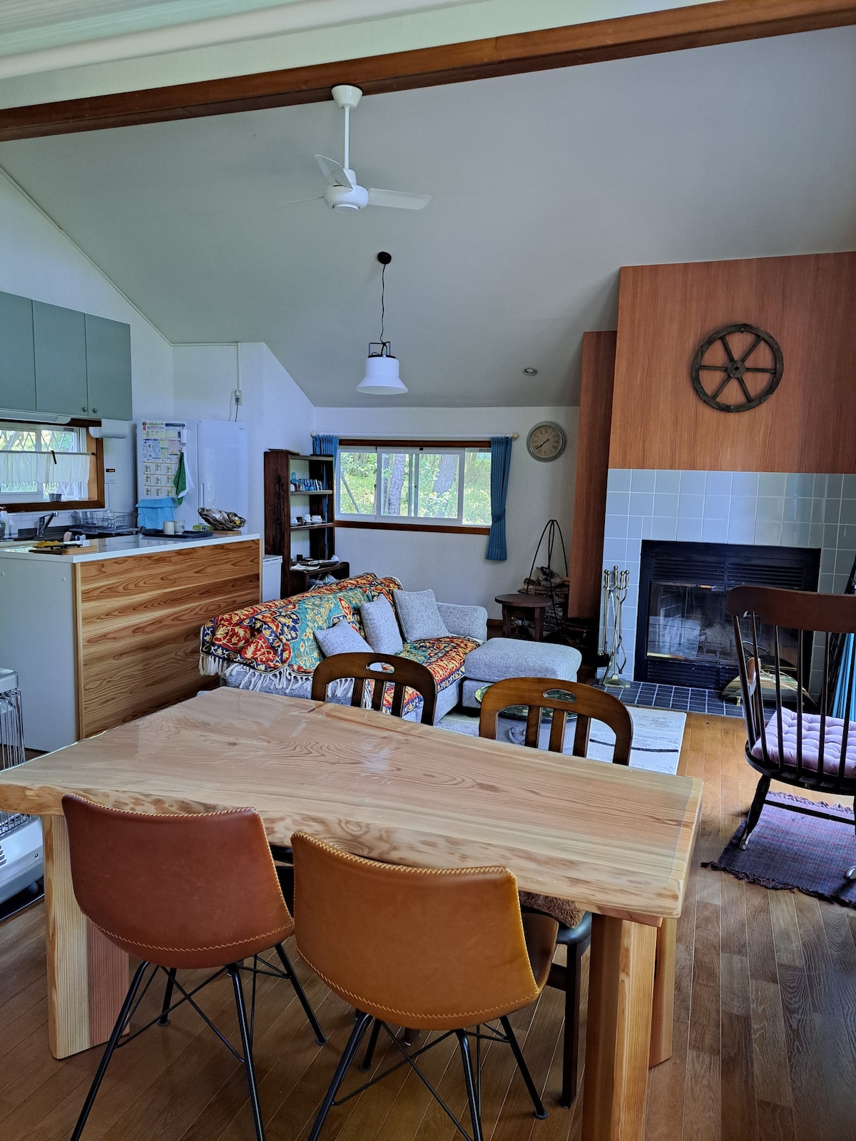 An open living space features a wooden dining table surrounded by four brown chairs. A cozy sofa with colorful cushions is positioned next to a fireplace, and large windows allow natural light to fill the room, enhancing the inviting environment.