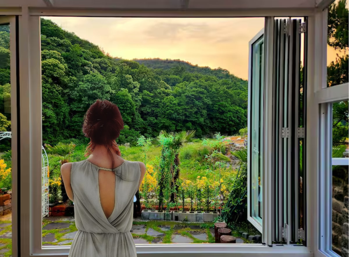 A view from an open window reveals a lush green landscape, framed by trees and illuminated by the soft glow of sunset. A person is seen from behind, looking out at the natural scenery, which includes patches of bright yellow flowers.