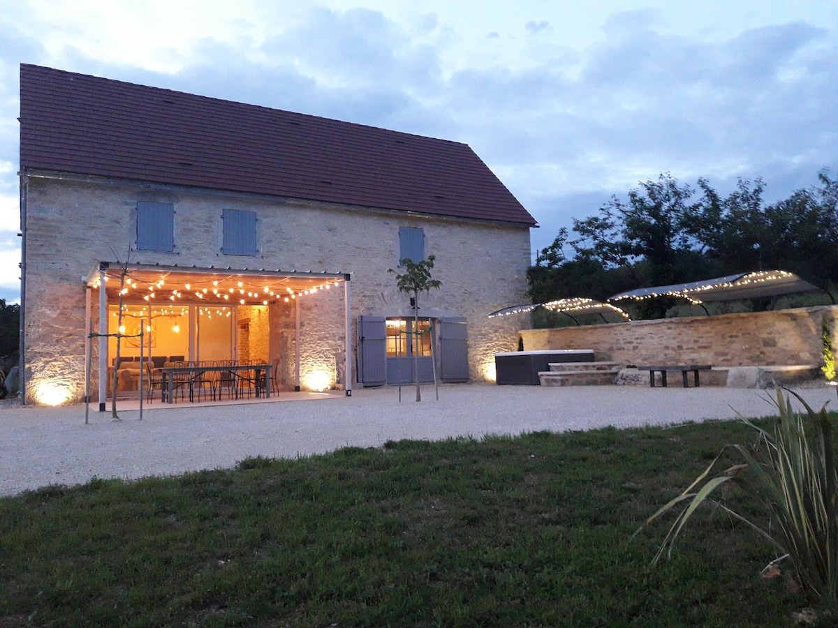 A spacious stone house is illuminated by soft outdoor lighting, showcasing a large terrace area with seating for gatherings. The exterior features blue shutters and a landscaped garden, providing a welcoming environment for relaxation and socializing.