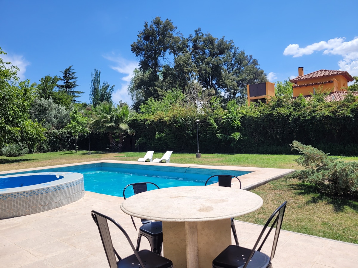 A sunny outdoor area features a pool surrounded by well-maintained grass and tall greenery. A circular table with black chairs is positioned in the foreground, while two lounge chairs are set near the pool. A jacuzzi is visible to the left.
