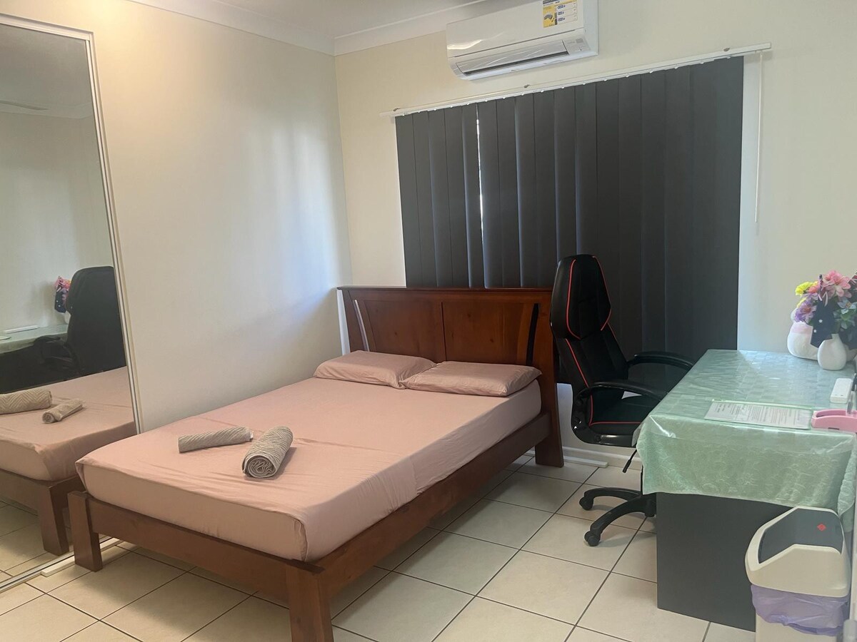 A furnished bedroom features a spacious bed covered with a light blanket and neatly rolled towels. A mirror reflects the room's layout. A desk and ergonomic chair are positioned near a window, while dark curtains provide privacy. The neutral-toned walls enhance the tranquil environment.