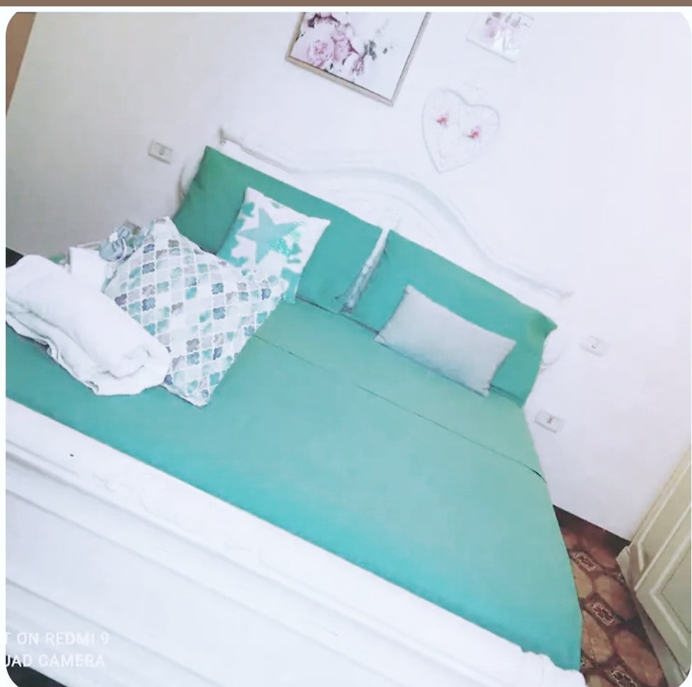 A cozy bedroom features a spacious bed with turquoise bedding, accented by decorative pillows in various patterns. Fresh white towels are neatly arranged on the side. Light-colored walls and a floral wall art add to the inviting atmosphere of the room.