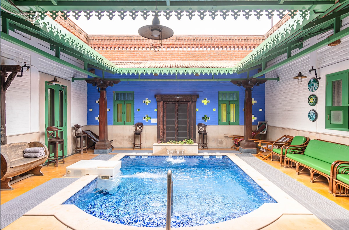 A tranquil courtyard features a sparkling blue pool at its center, framed by vibrant green accents and ornate architectural details. Comfortable seating options are provided alongside the pool, with traditional wooden elements and decorative wall art enhancing the serene atmosphere.