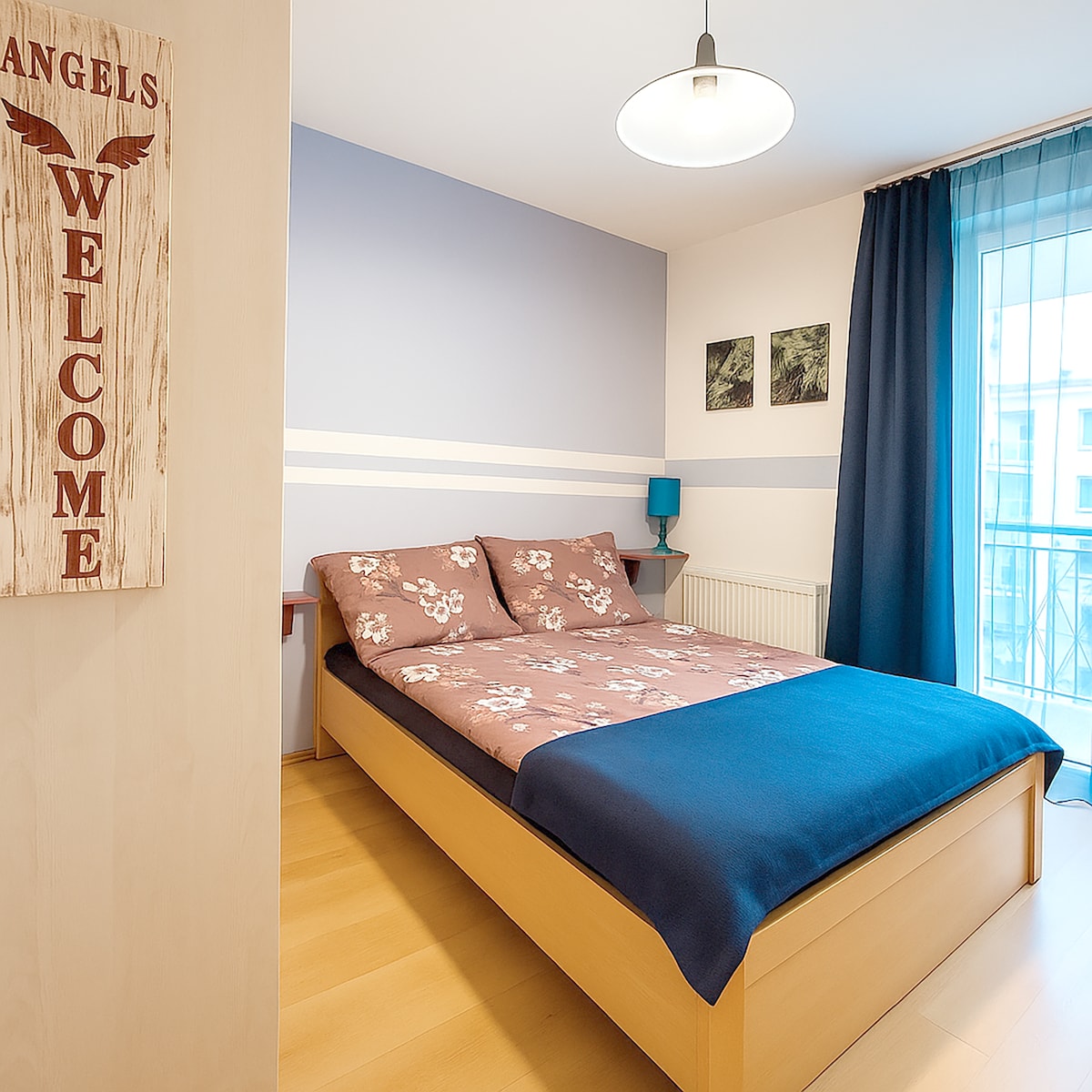 A well-lit bedroom features a spacious bed with a floral patterned cover and a solid blue throw. Light blue walls are complemented by dark curtains, while framed artwork adorns one side. A small table and lamp add to the room's functionality and comfort.