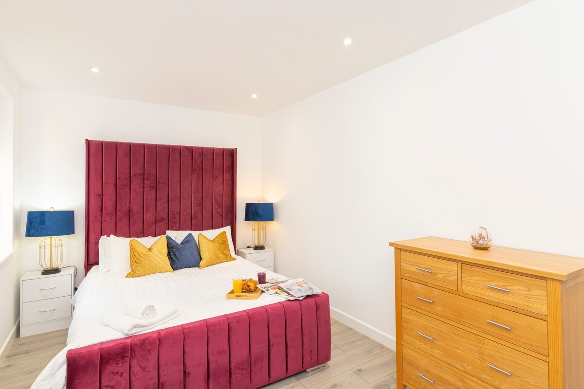 A spacious bedroom features a plush bed with an upholstered headboard in deep burgundy. Bright yellow and blue accent pillows are arranged on the bed. Nightstands with lamps flank the bed, and a wooden dresser provides additional storage against the white wall.