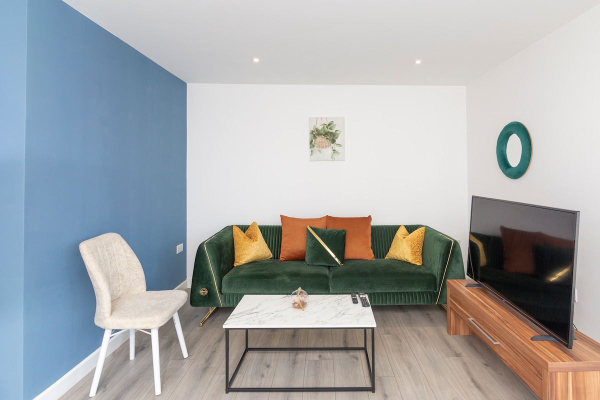 A modern living area features a green velvet sofa adorned with orange and gold cushions, providing a welcoming seating space. A sleek coffee table sits in front, complemented by a light wooden television unit. A simple wall decoration and round mirror add subtle details to the room.