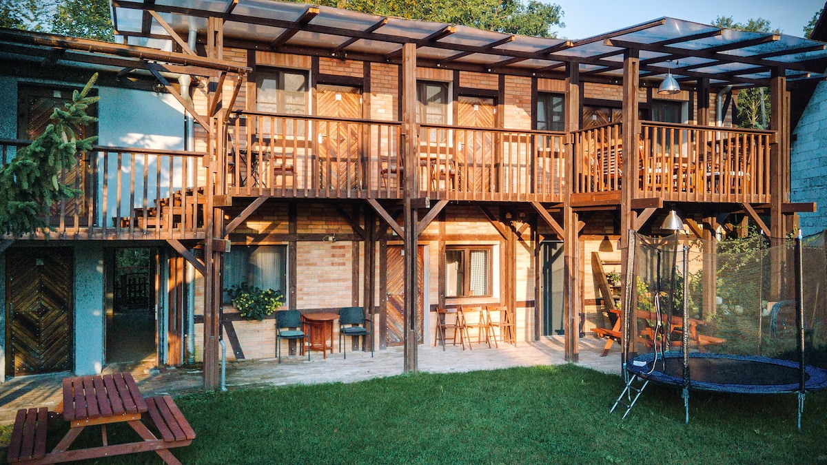 The exterior of a multi-level wooden lodge is showcased, featuring balconies with seating and vibrant green grass in the foreground. The structure is supported by sturdy beams, with a trampoline and picnic table visible in the outdoor area.