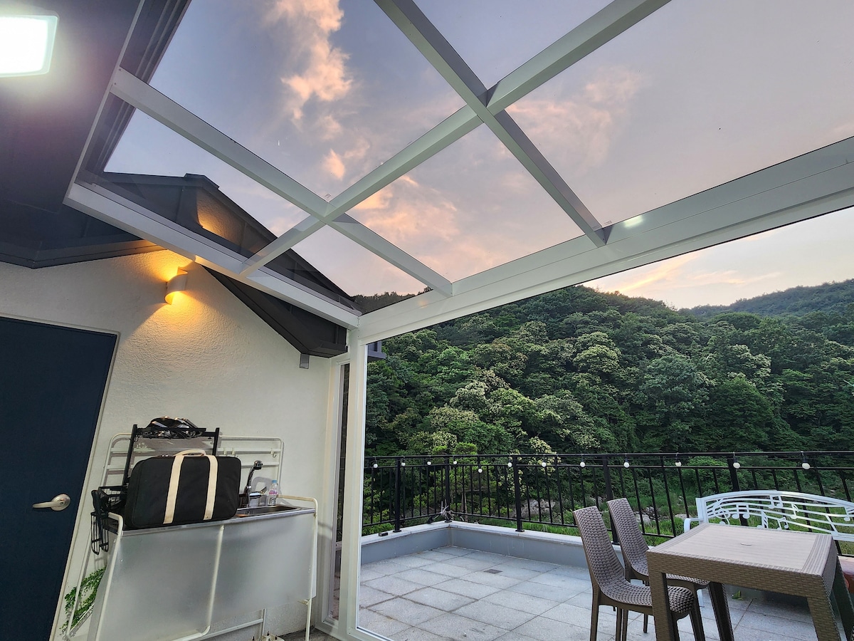 A spacious terrace is shown, featuring a dining table with several chairs set against a backdrop of lush greenery. A small sink and storage area are visible, while large glass panels allow ample natural light. The sky exhibits soft colors of dusk.