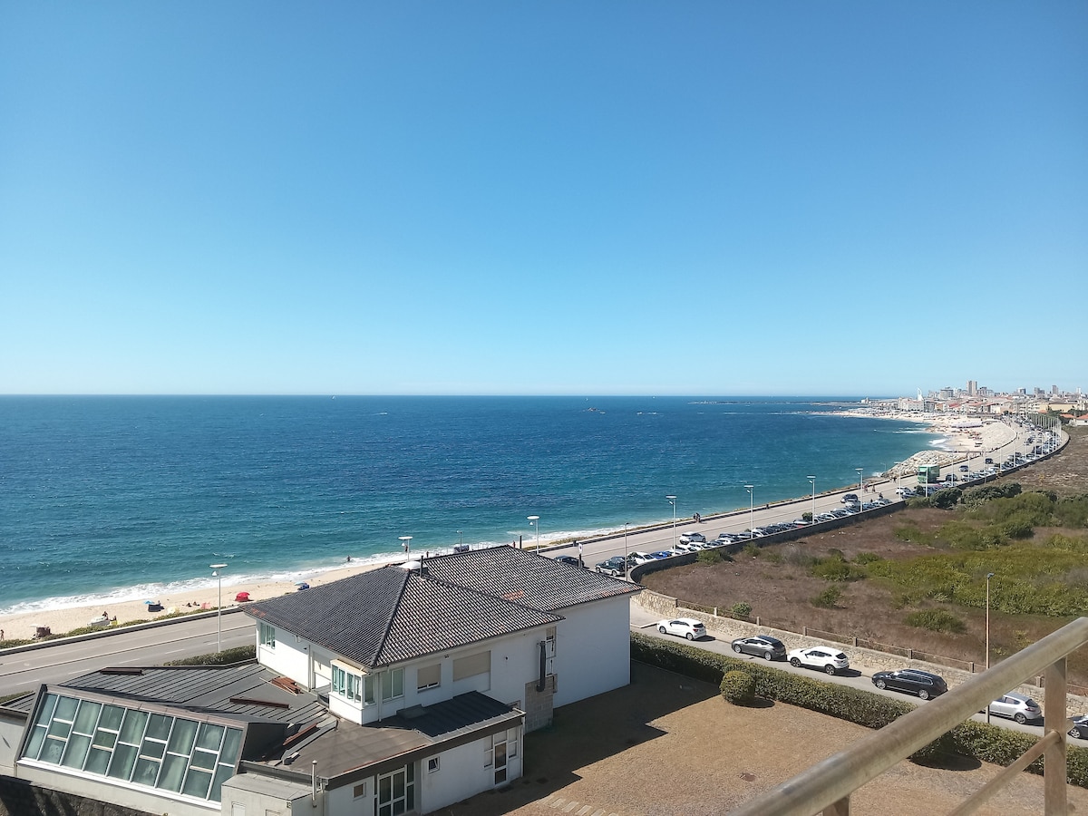 A sweeping coastal view reveals a sandy beach bordered by a gentle wave-lapped shoreline. In the foreground, a modern building can be seen, with a backdrop of clear blue skies and distant cityscapes along the horizon.