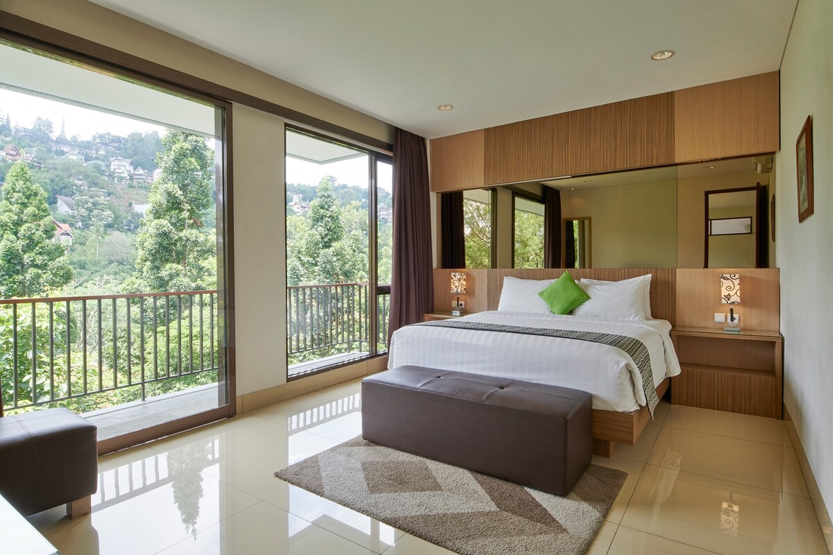 A spacious bedroom is highlighted by large windows offering hillside and forest views. A comfortable bed is centered against a wooden wall, adorned with a green accent pillow. A bench sits at the foot of the bed, and natural light fills the room.