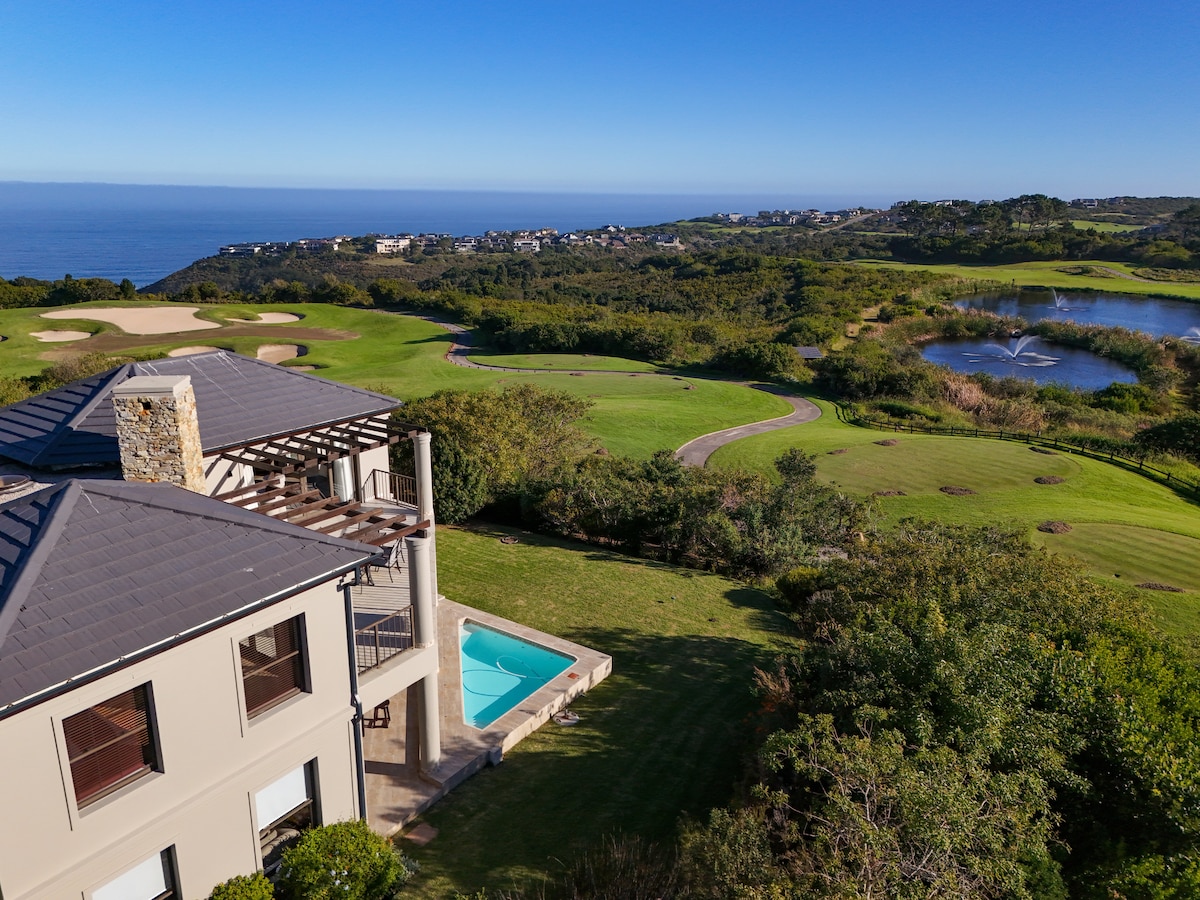 An aerial view showcases the Pezula Lake House, featuring a private splash pool adjacent to the open lawn. The surrounding green landscape extends to the golf course, blending with scenic views of the ocean in the background. Natural vegetation enhances the serene environment.