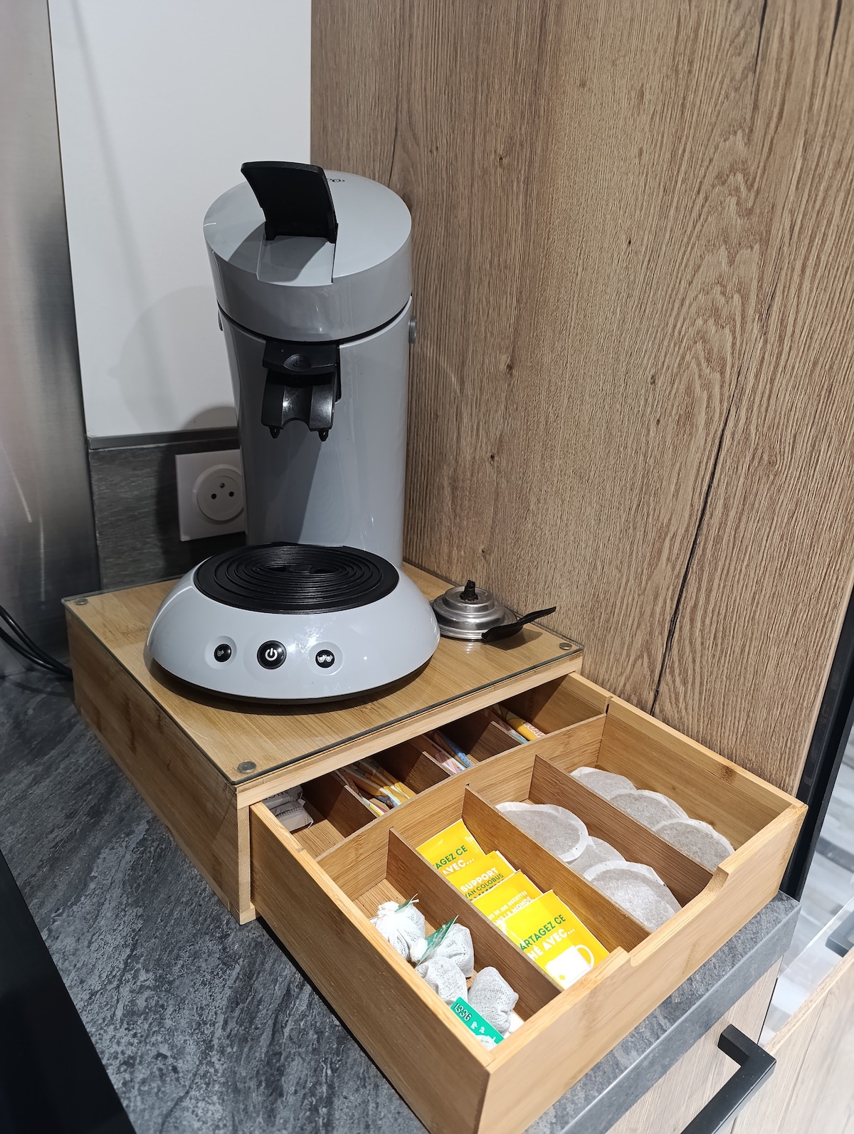 A coffee maker with a sleek gray design is positioned atop a wooden tray. Below, a bamboo drawer contains individual compartments filled with tea bags, sugar packets, and coffee pods, offering a convenient selection for guests.