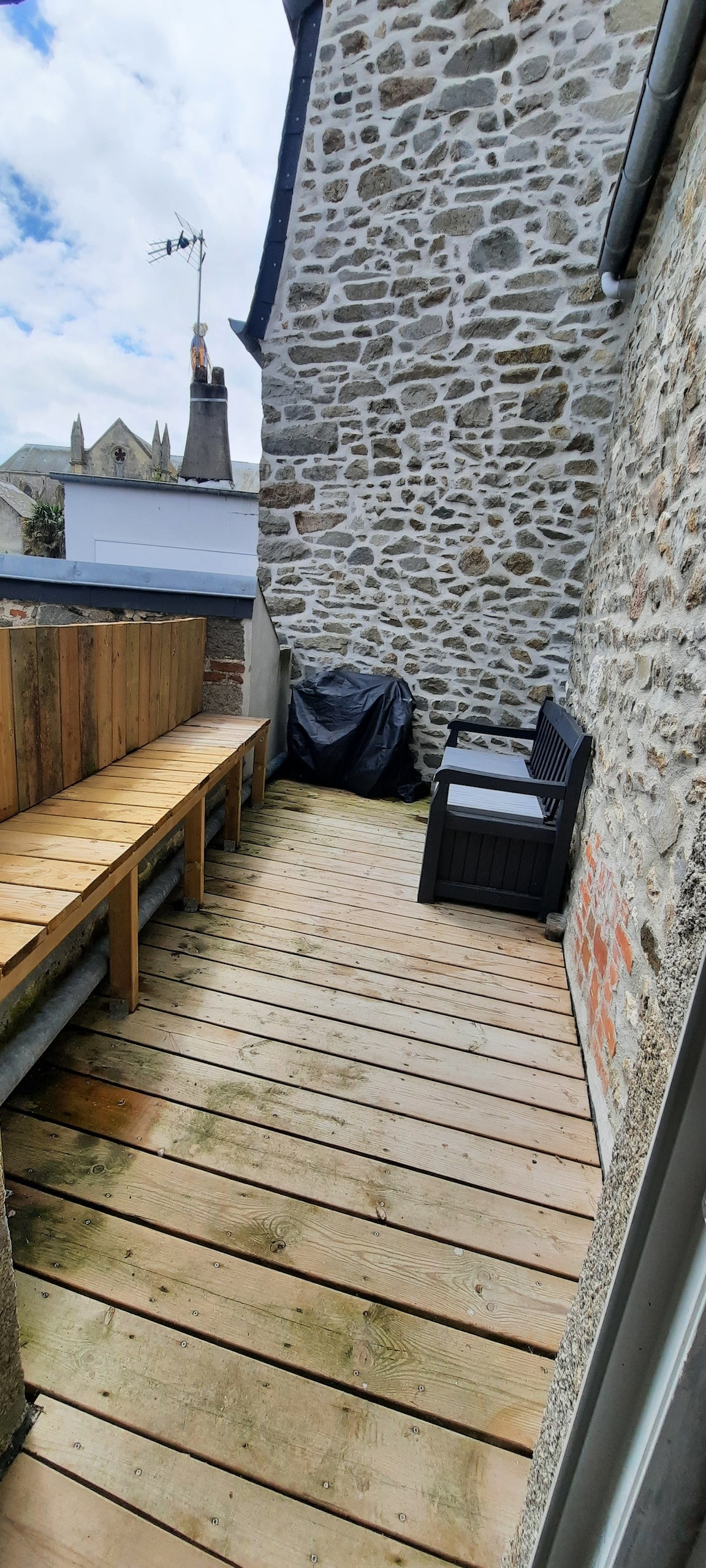 A wooden terrace area is revealed, featuring a wooden railing and a small seating arrangement with two chairs. The space is bordered by stone walls and offers a view of neighboring buildings. A barbecue is positioned in one corner, enhancing outdoor possibilities.