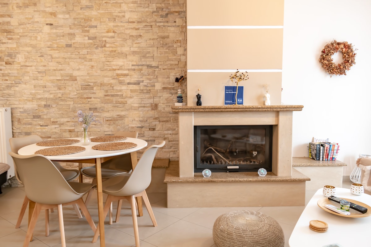 The living area showcases a modern dining table with four light-colored chairs, positioned near a stone feature wall. A decorative fireplace is integrated into the space, adding to its cozy ambiance. A small decorative wreath is displayed above, enhancing the room's design.