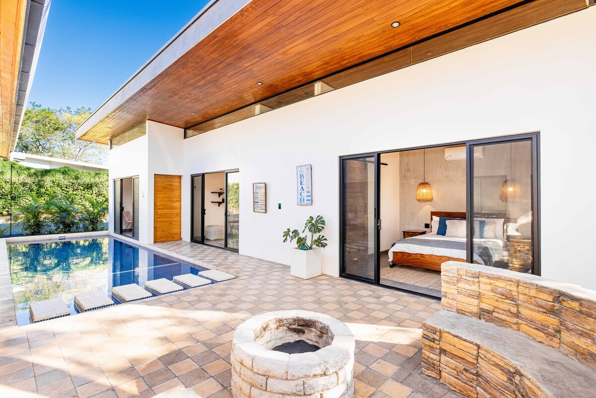 An open-air area features a private pool bordered by elegant tiles. On one side, a comfortable sleeping space is visible through glass doors, while a stone seating area encircles a fire pit. Lush greenery is present in the background, enhancing the tranquil atmosphere.