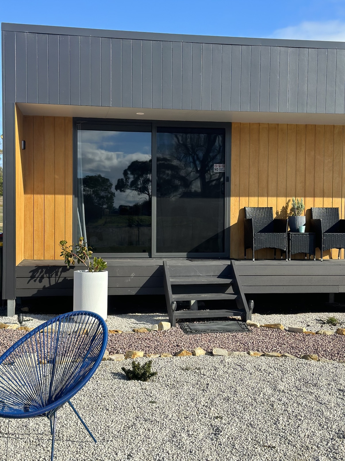 The cabin exterior is designed with a mix of timber and modern materials, featuring a large sliding glass door. A small deck is accessed by steps, with two stylish outdoor chairs positioned nearby. Decorative plants and gravel landscaping enhance the inviting entrance.