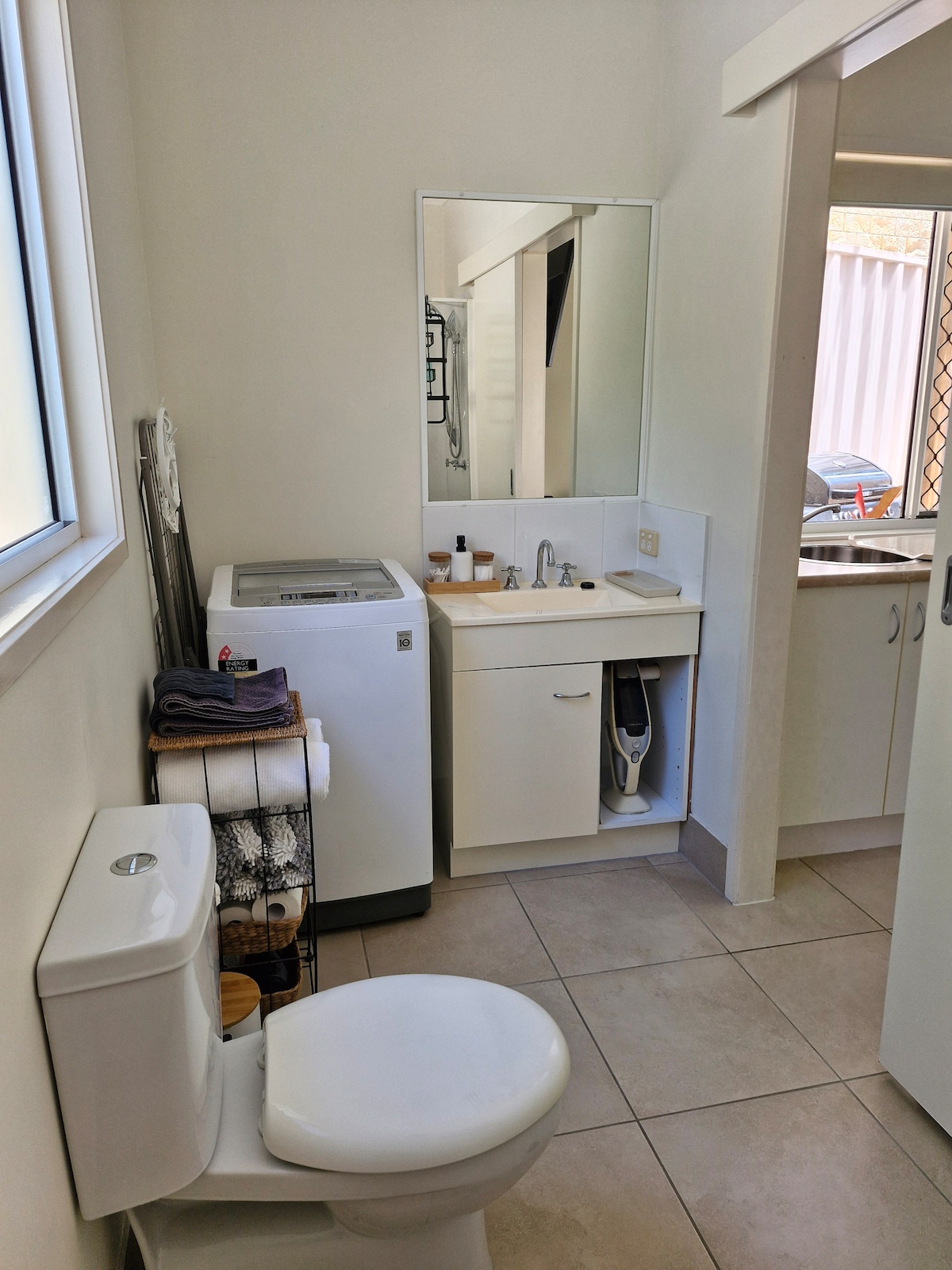 A clean bathroom is shown, equipped with a toilet, washing machine, and a sink. A mirror reflects the space, while towels are neatly arranged on a rack. Natural light from a window adds brightness to the light-colored fixtures and tiled flooring.