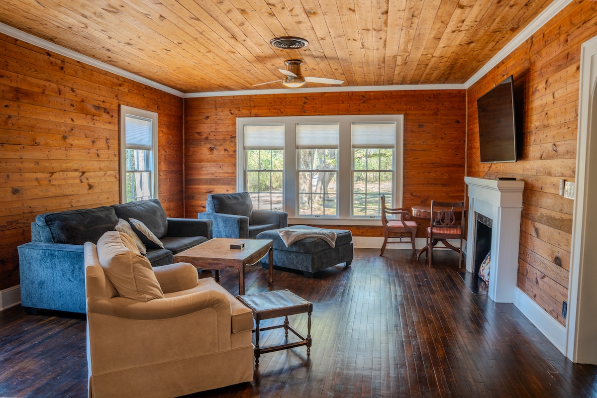 A spacious living area features warm wooden walls and a ceiling, with large windows allowing natural light. Comfortable seating includes a sectional sofa, armchair, and a coffee table in the center. A flat-screen TV is mounted on the wall near the corner.