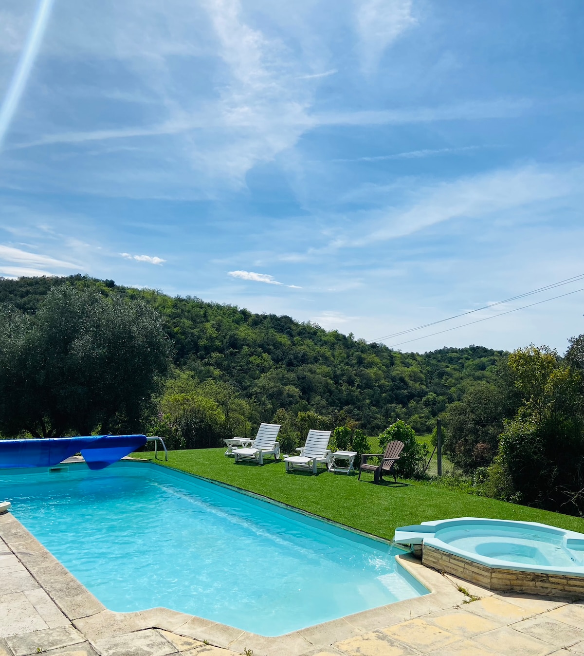 A serene outdoor area features a refreshing swimming pool surrounded by lush grass, with several lounge chairs positioned for relaxation. Nearby, a jacuzzi invites guests to unwind, all set against a backdrop of gently rolling green hills under a clear blue sky.