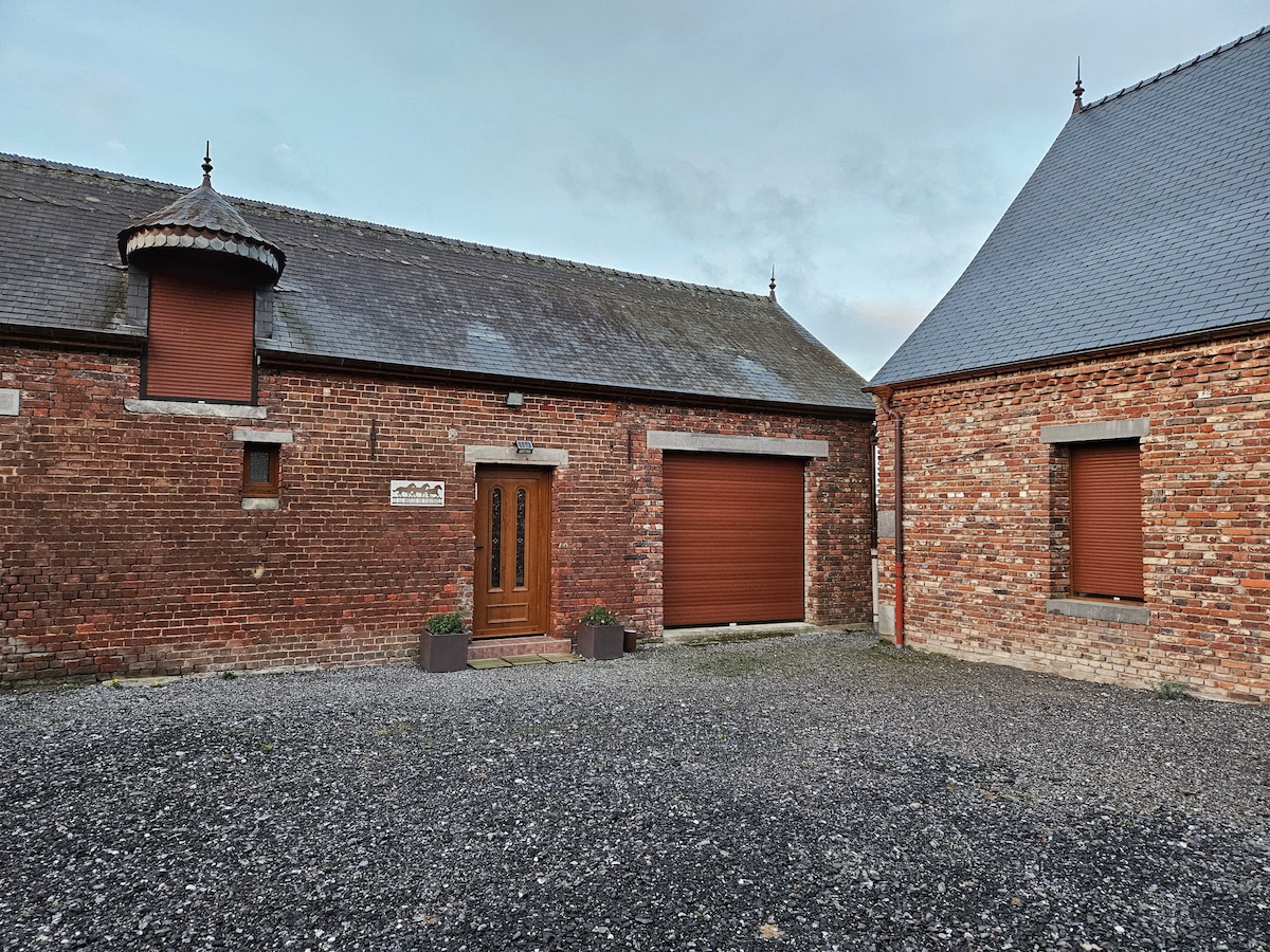 The exterior of the accommodation is shown, featuring a brick facade with a distinct architectural design. A central entrance with double doors is flanked by potted plants. The surrounding gravel area adds to the inviting atmosphere, while the neighboring structure exhibits a complementary design.