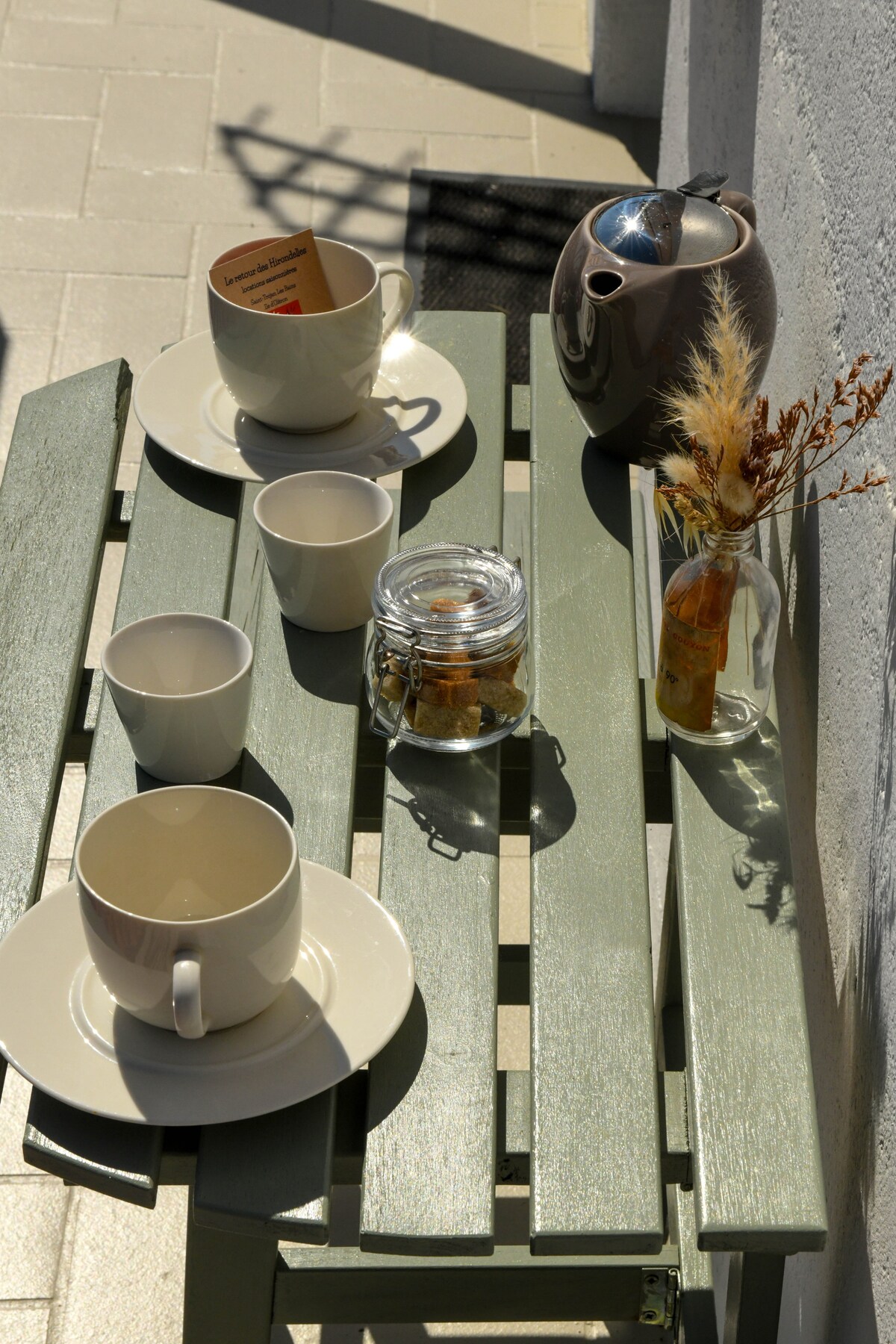 A small outdoor table is set for coffee or breakfast. Two larger cups and three smaller ones rest on beige plates, accompanied by a glass jar of cookies and a slender vase containing dried flowers. Soft sunlight illuminates the scene.