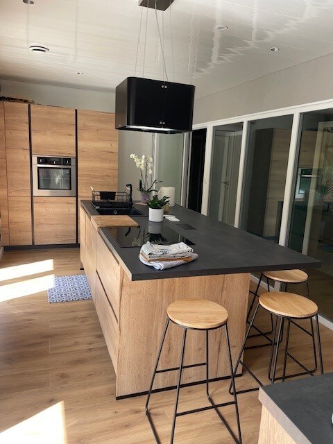 A modern kitchen features wooden cabinetry and sleek black countertops. A central island with two metal bar stools invites casual seating. Light reflects off the surfaces, and a vase with an orchid adds a touch of greenery. Large glass doors provide a connection to the outdoor area.