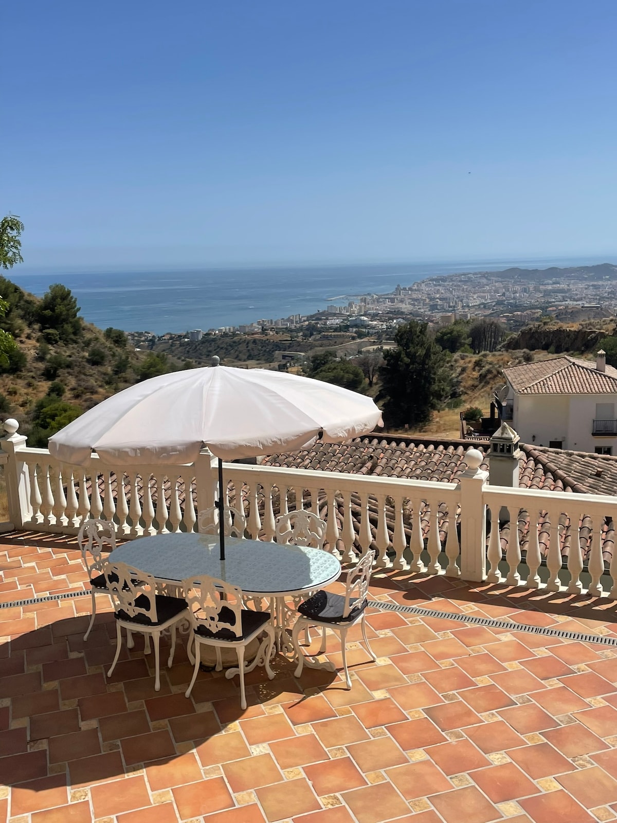 A round dining table with elegant white chairs is set under a large umbrella on a tiled terrace. The view encompasses a serene coastline and rolling hills, with a clear blue sky overhead.