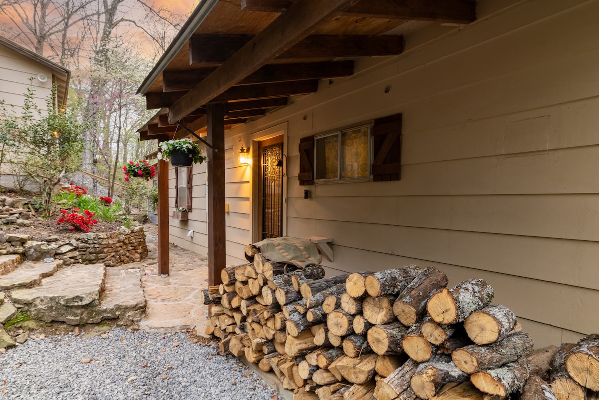 A quaint cottage exterior showcases a stacked pile of logs, ready for use by the cozy fireplace indoors. Flower pots hang above the door, adding a touch of greenery. The stone pathway leads to a welcoming entrance, surrounded by garden beds with seasonal blooms.