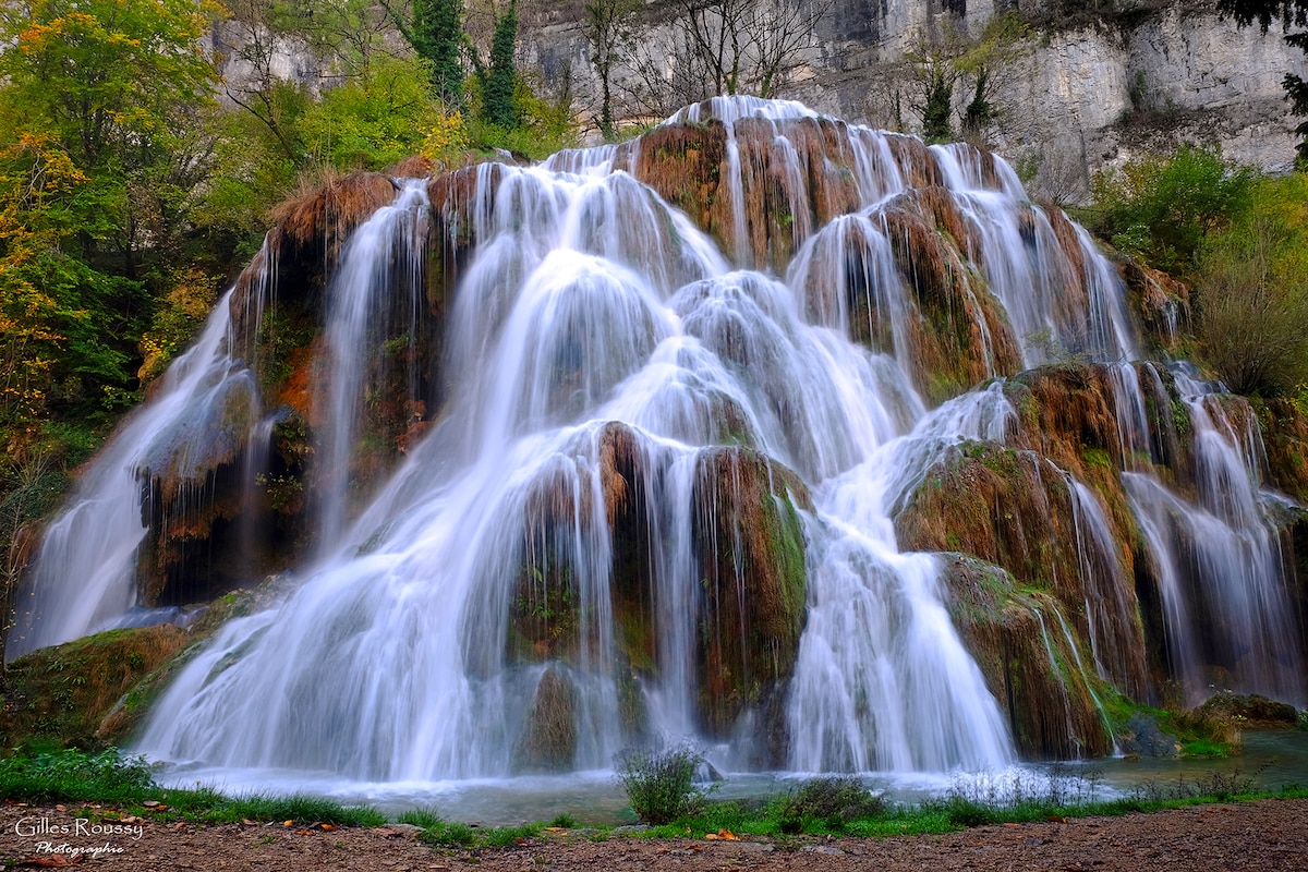 A cascading waterfall flows over a rocky landscape, surrounded by greenery and autumn foliage. The water appears silky as it tumbles down multiple tiers, pooling at the base. The scene conveys a sense of peacefulness and natural beauty.