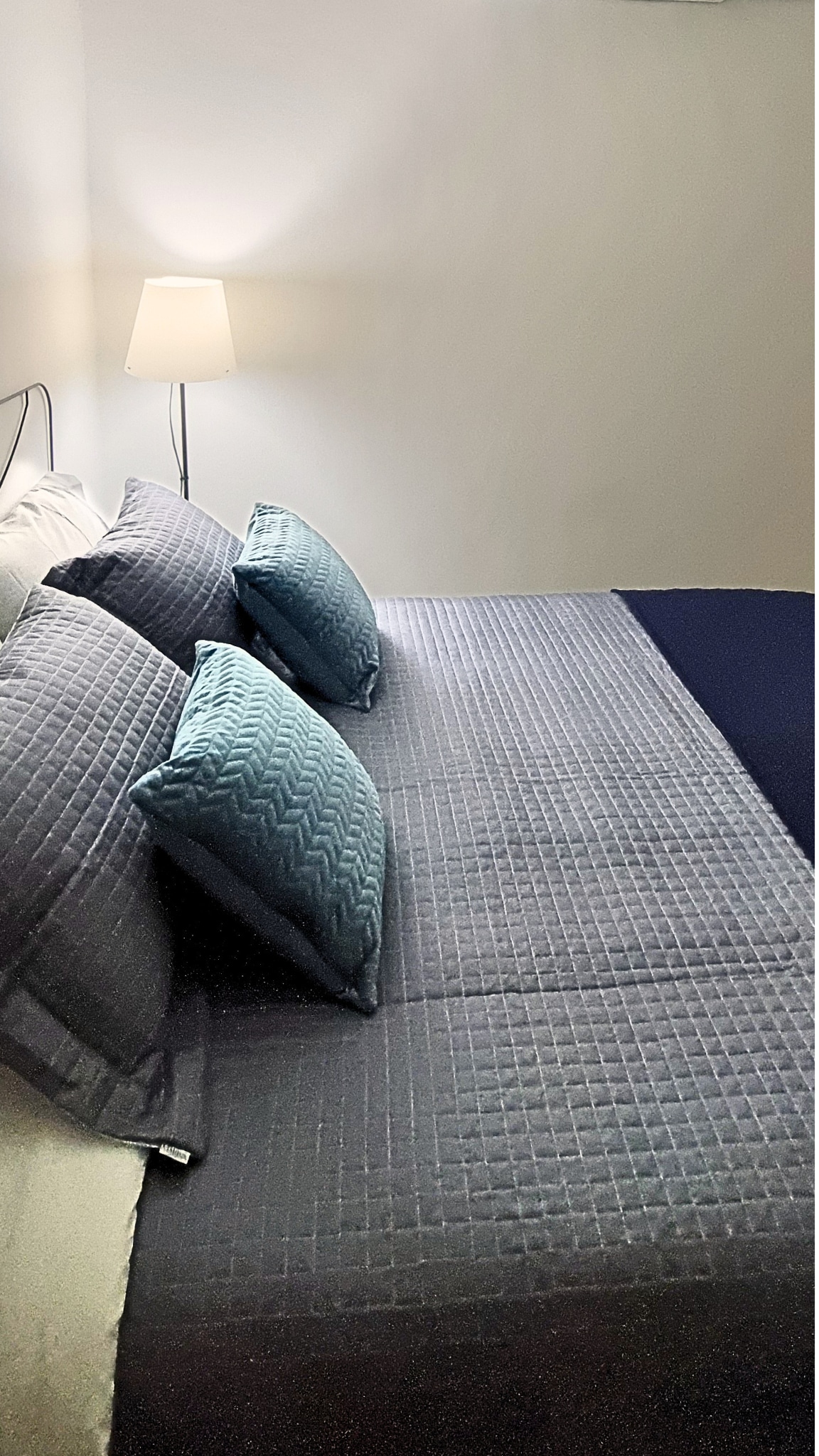 A well-arranged sleeping area is presented, featuring a spacious bed covered with a textured gray bedspread. Decorative pillows in varying shades of blue are placed neatly on the bed. A soft glow is emitted from a nearby lamp, providing gentle illumination in the room.