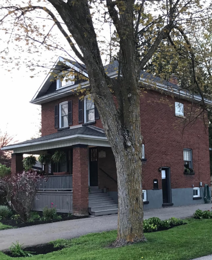 A charming three-story brick home stands on a grassy front yard, framed by a large tree. The porch is accessible through steps leading to the main entrance. Greenery and flowerbeds enhance the exterior, showcasing a welcoming presence on the quiet residential street.