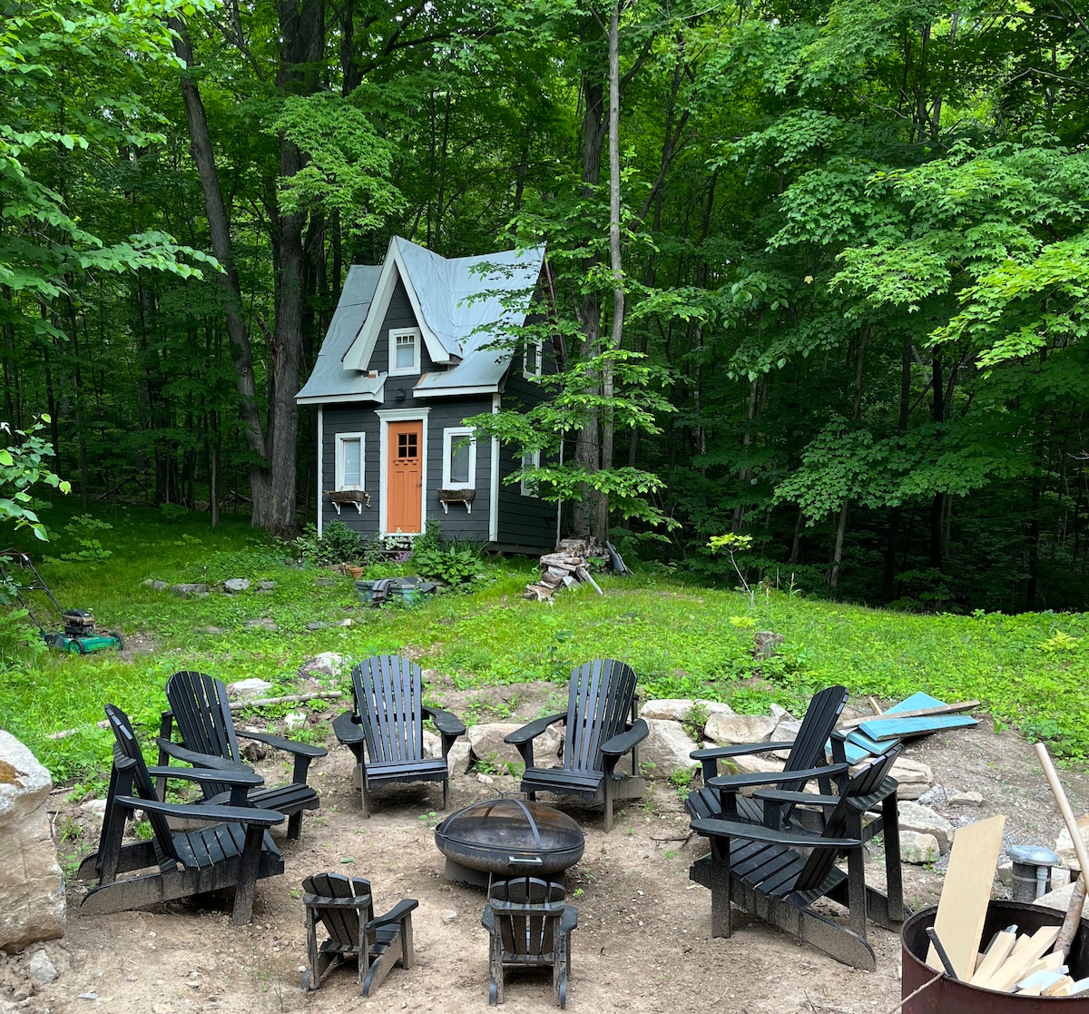 A charming cabin is surrounded by lush greenery, featuring a welcoming entrance with an orange door. Around a circular fire pit, multiple black Adirondack chairs are arranged on a natural dirt surface, providing a cozy gathering space in a serene woodland setting.