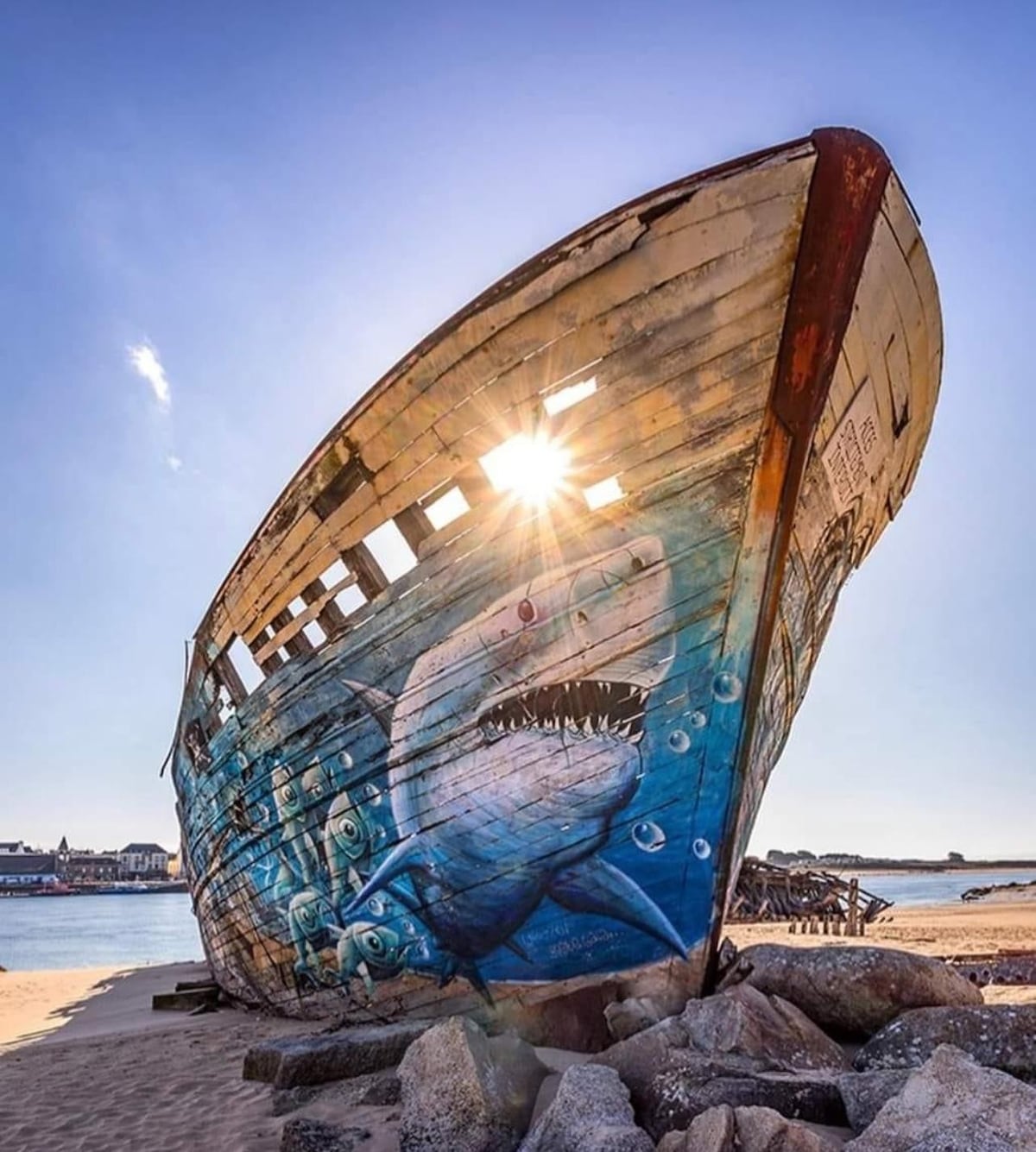 An old wooden boat stands on a sandy beach, featuring a colorful mural of a shark. The sun shines through the boat, creating a halo effect. In the background, beach houses and rocky formations are visible against a clear blue sky.