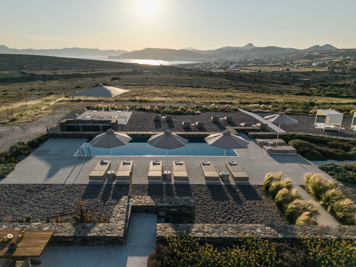 An expansive outdoor area features a pool surrounded by sun loungers and umbrellas, with a gently landscaped garden adjoining the space. The horizon showcases distant hills and the shimmering sea under a soft evening light.