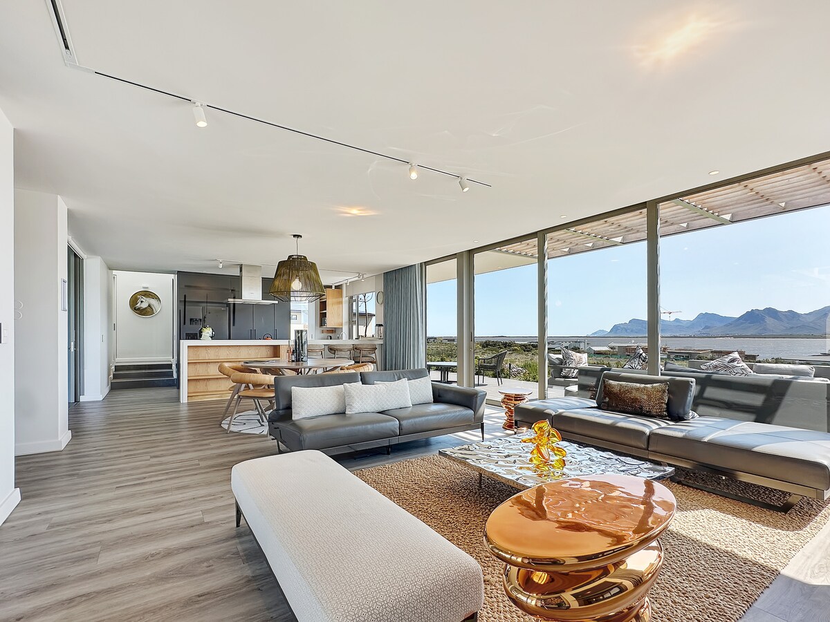 A spacious living area features sleek furniture arranged thoughtfully. Large windows provide panoramic views of the surrounding landscape, while a stylish coffee table reflects light. A dining area and modern kitchen are seen in the background, enhancing the open-concept design.