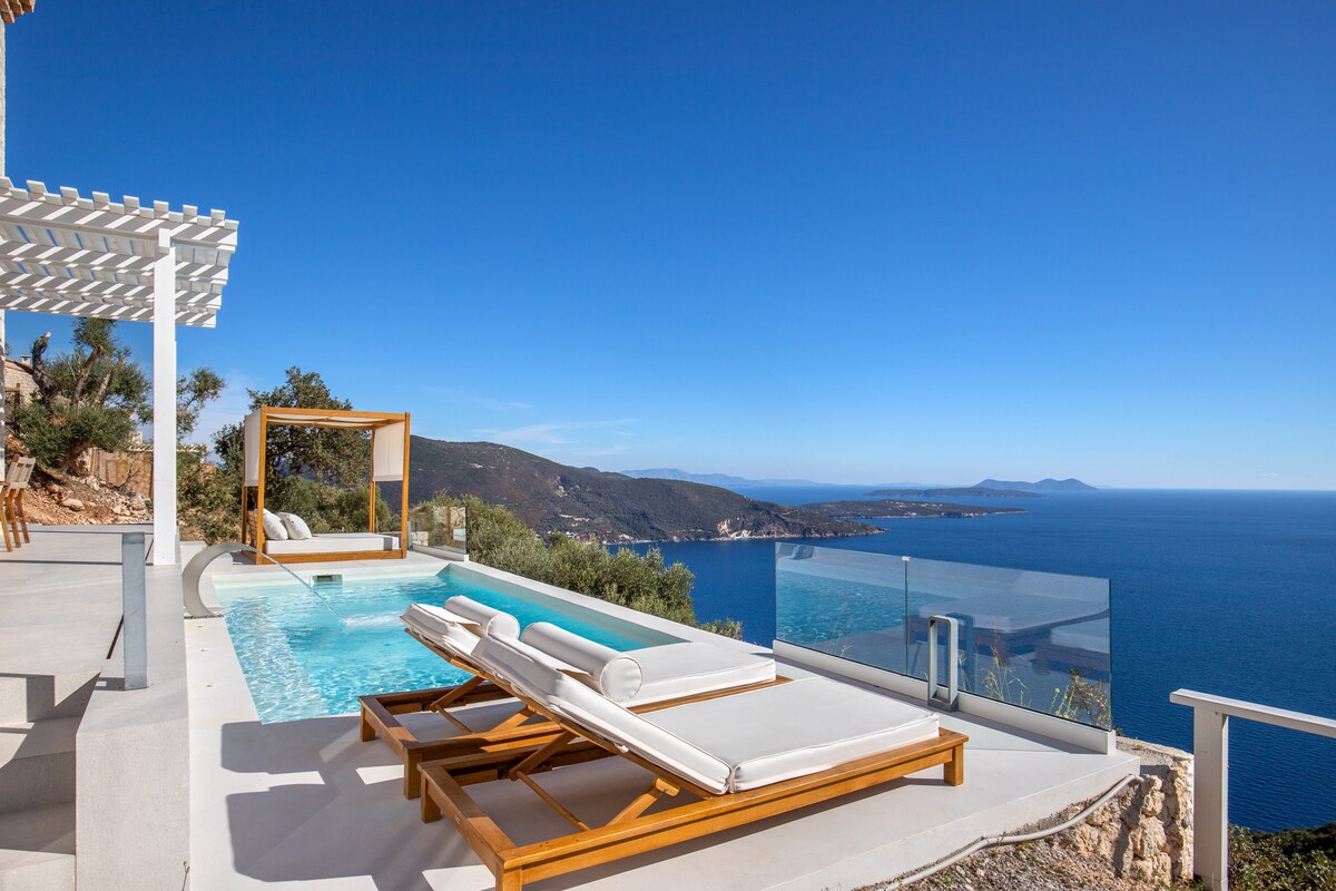 A serene outdoor space features a saltwater pool with stunning ocean views. Sun loungers are arranged next to the pool, providing a perfect spot for relaxation. A glass fence offers unobstructed views of the neighboring islands against a clear blue sky.