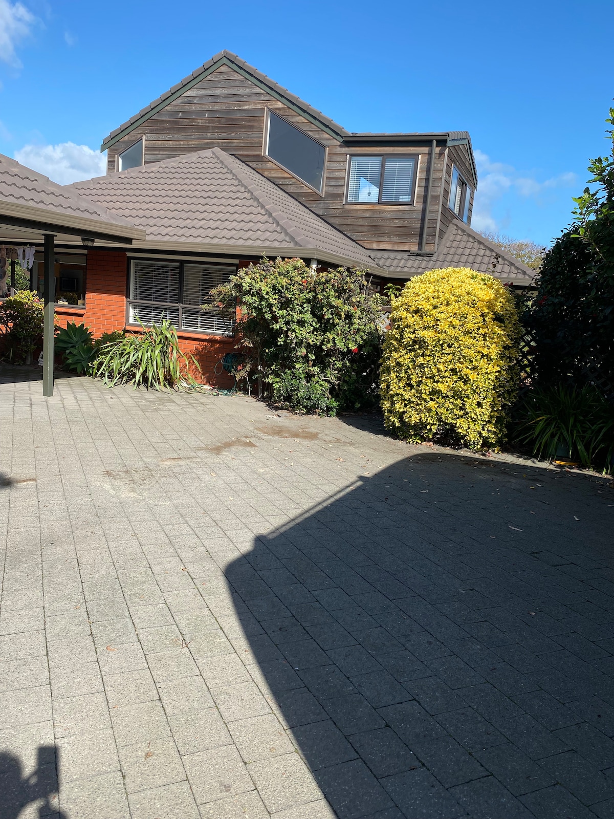 The exterior view of a two-story family home is presented, featuring a mix of brick and wooden finishes. Lush greenery, including well-maintained shrubs and trees, borders the paved driveway. The clear blue sky enhances the inviting atmosphere of the property.