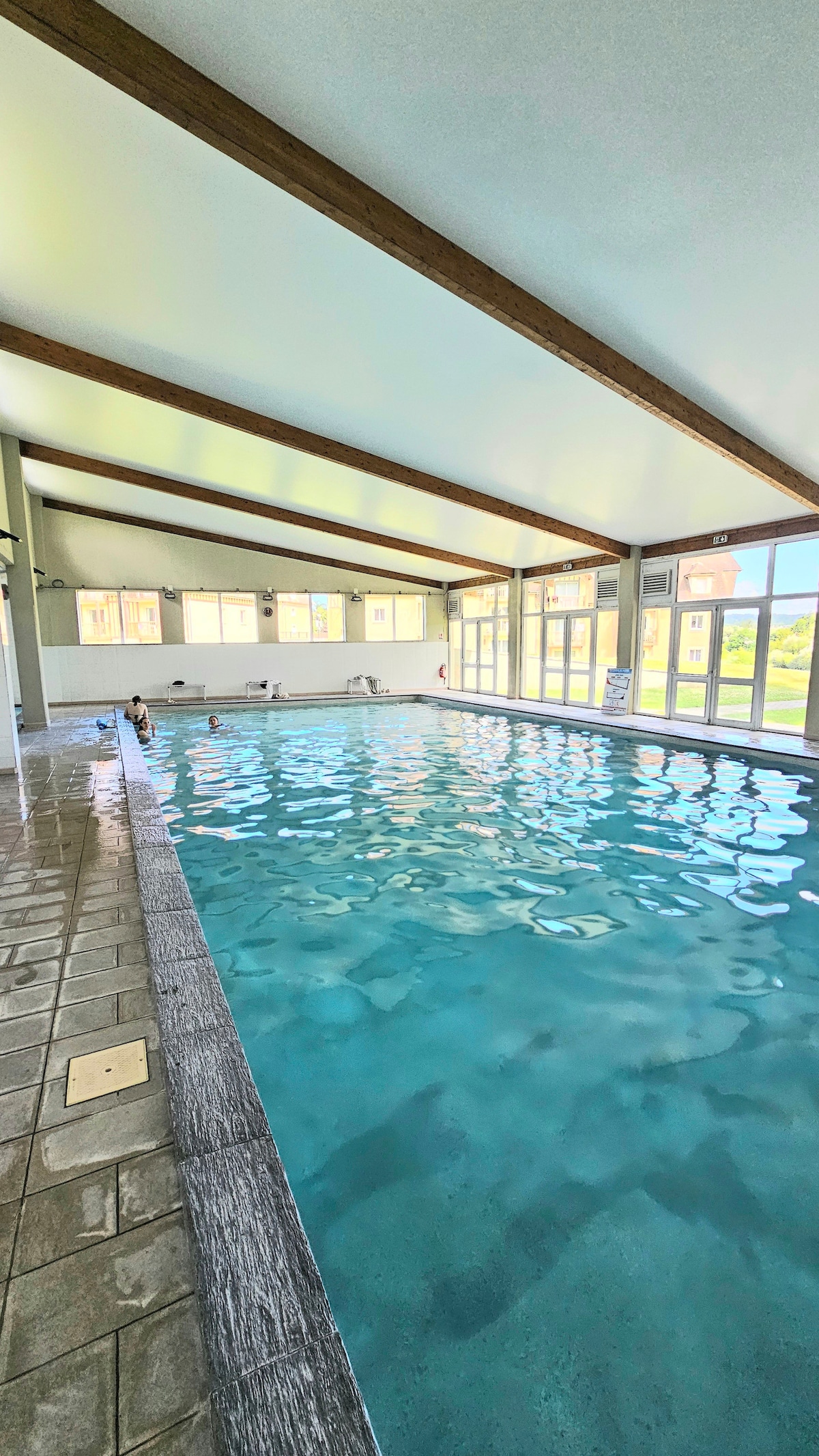 An indoor swimming pool is highlighted by large windows that allow natural light to fill the space. The pool features a clean, blue surface and is bordered by tiled flooring. Wooden beams add architectural interest to the ceiling, enhancing the spacious feel of the area.