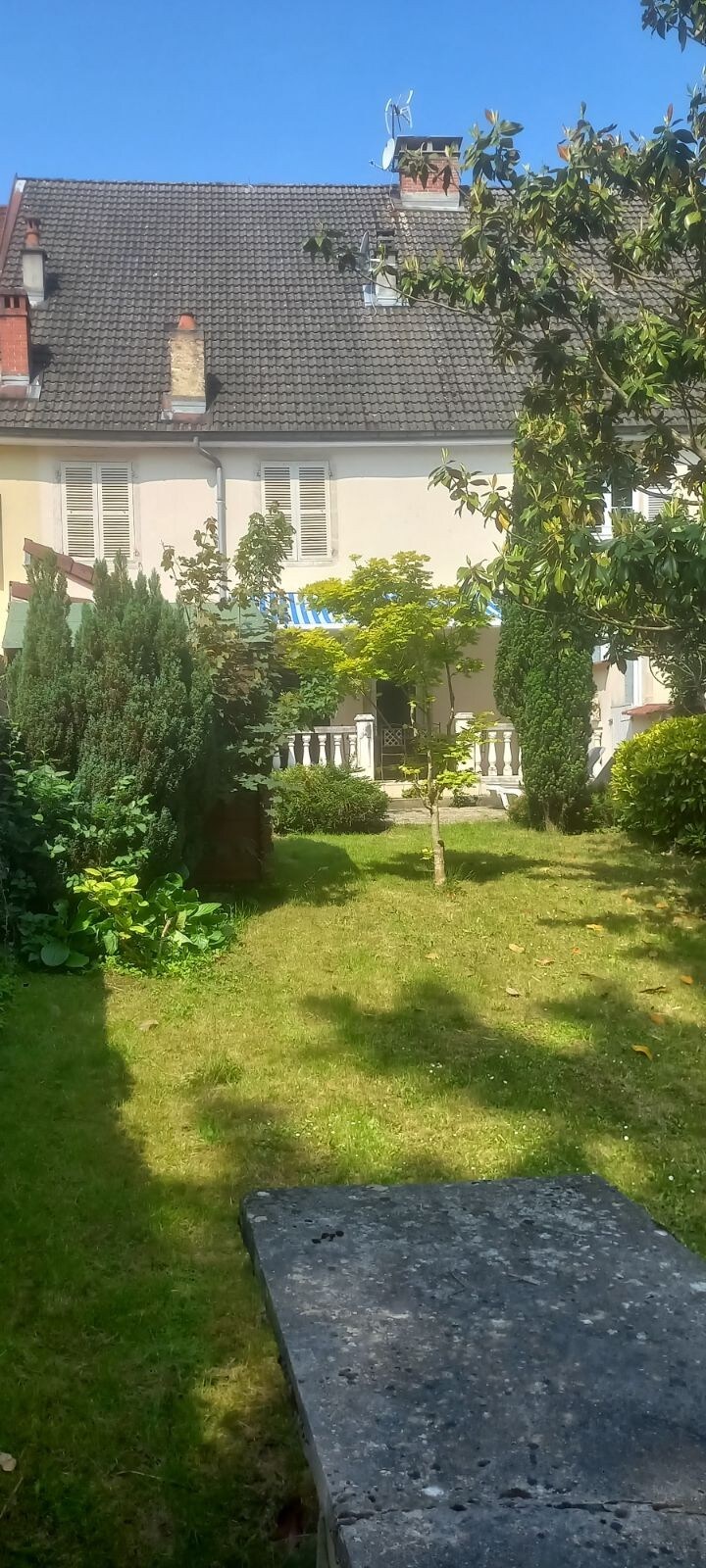 The garden area features a well-maintained lawn surrounded by various shrubs and trees. A stone pathway leads to a charming house, which has a classic exterior and bright awnings. The clear sky enhances the inviting atmosphere of this peaceful outdoor space.