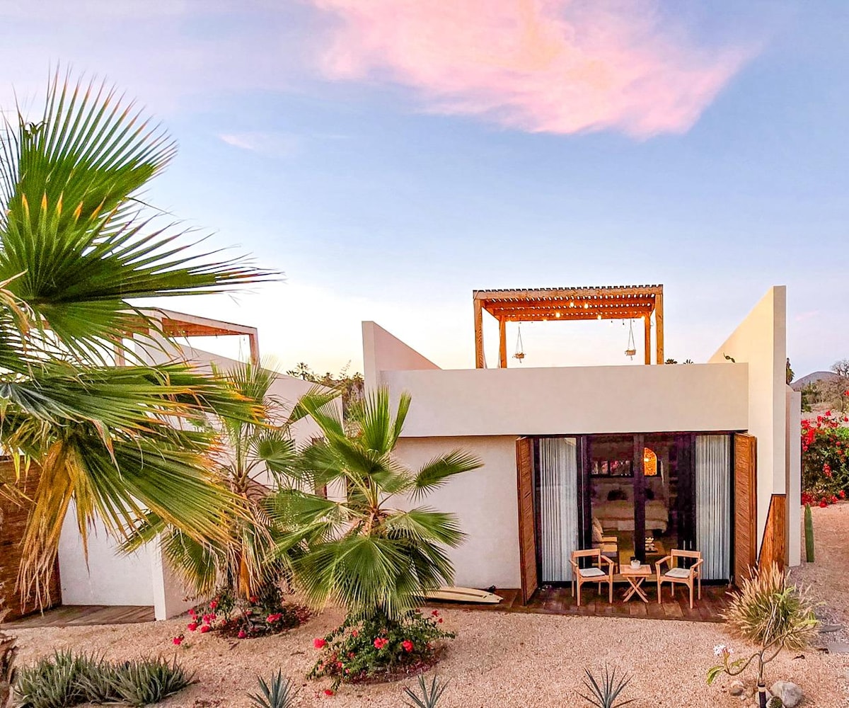 The exterior of the bungalow showcases modern architecture surrounded by desert flora. A wooden pergola extends above the front porch, where a small dining set is visible. A backdrop of vibrant bougainvillea adds a touch of color against a pastel sky.