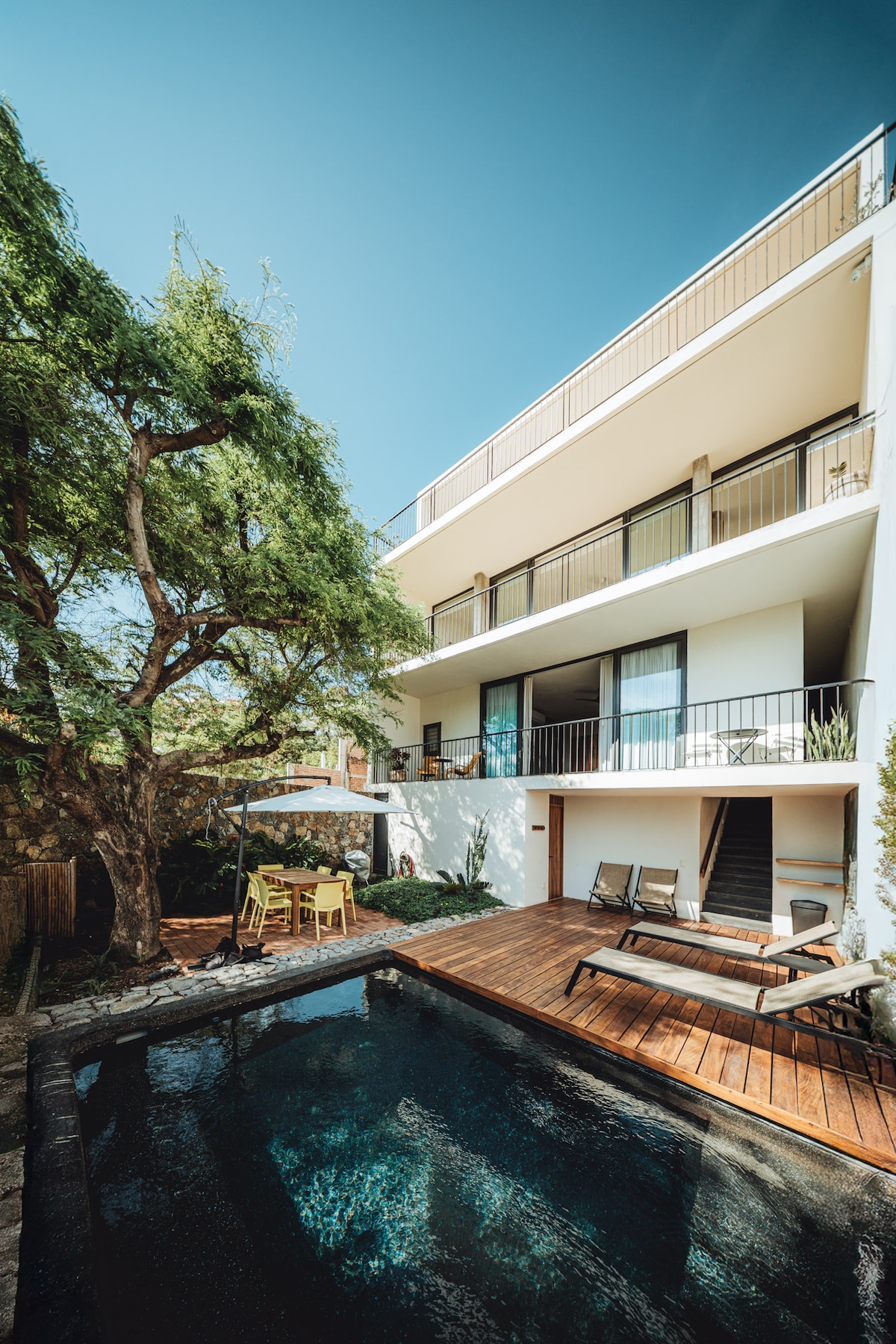 The exterior view of the house reveals a modern design with multiple levels. A spacious pool is surrounded by wooden decking and lounge chairs. Lush greenery, including a sizable tree, enhances the outdoor space. Balconies are seen from the upper floors, offering additional seating.